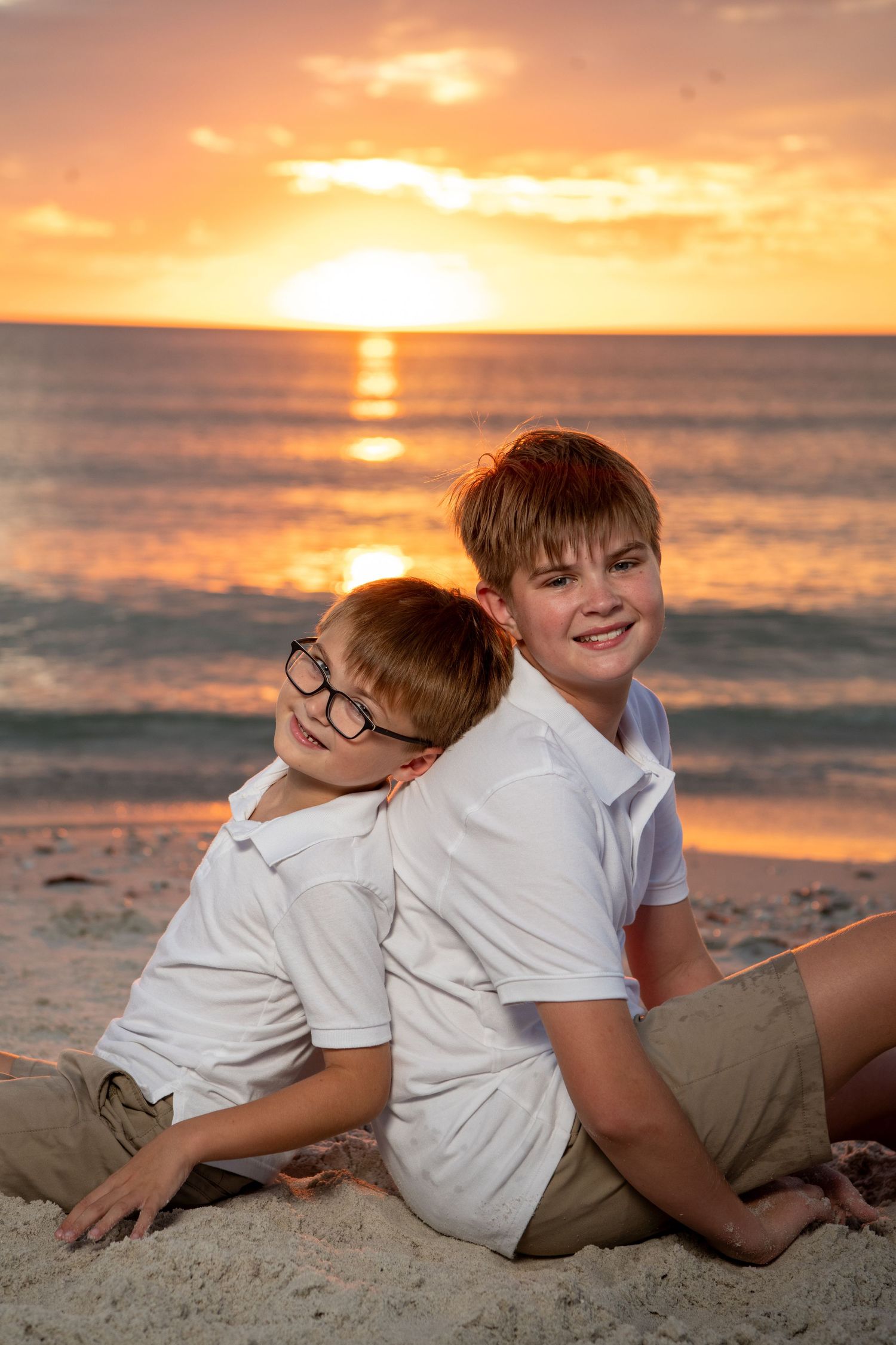 Two young people in white shirts sitting on a beach at sunset with golden orange sky and ocean waves. Beach photoshoot. Family photoshoot,