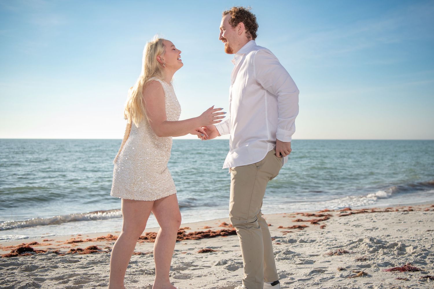 A romantic beach photoshoot featuring a couple in white outfits against a bright blue ocean backdrop. Engagement photoshoot. Couples photos.
