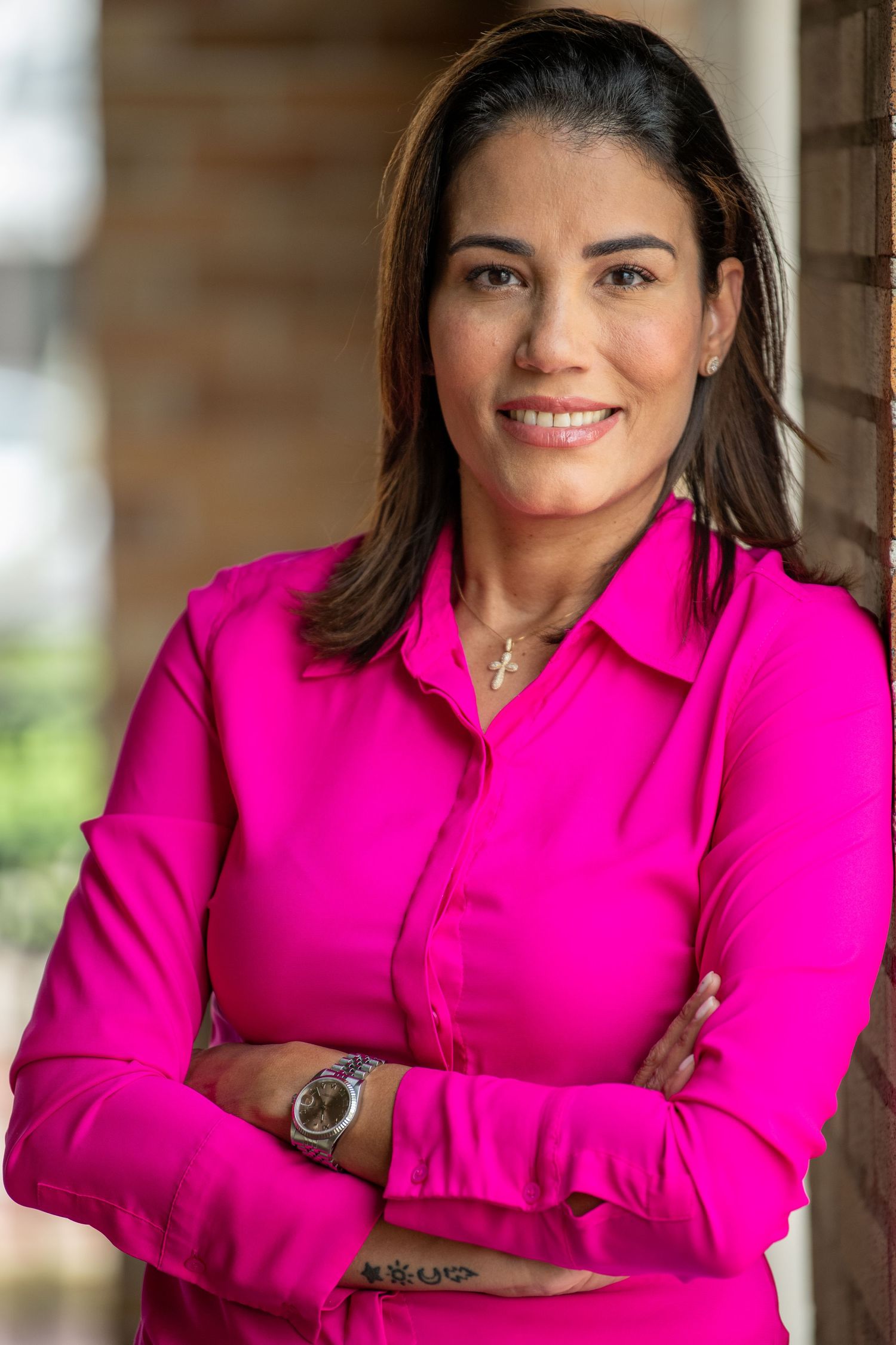 Professional portrait of person in vibrant pink blouse with jewelry against brick background.