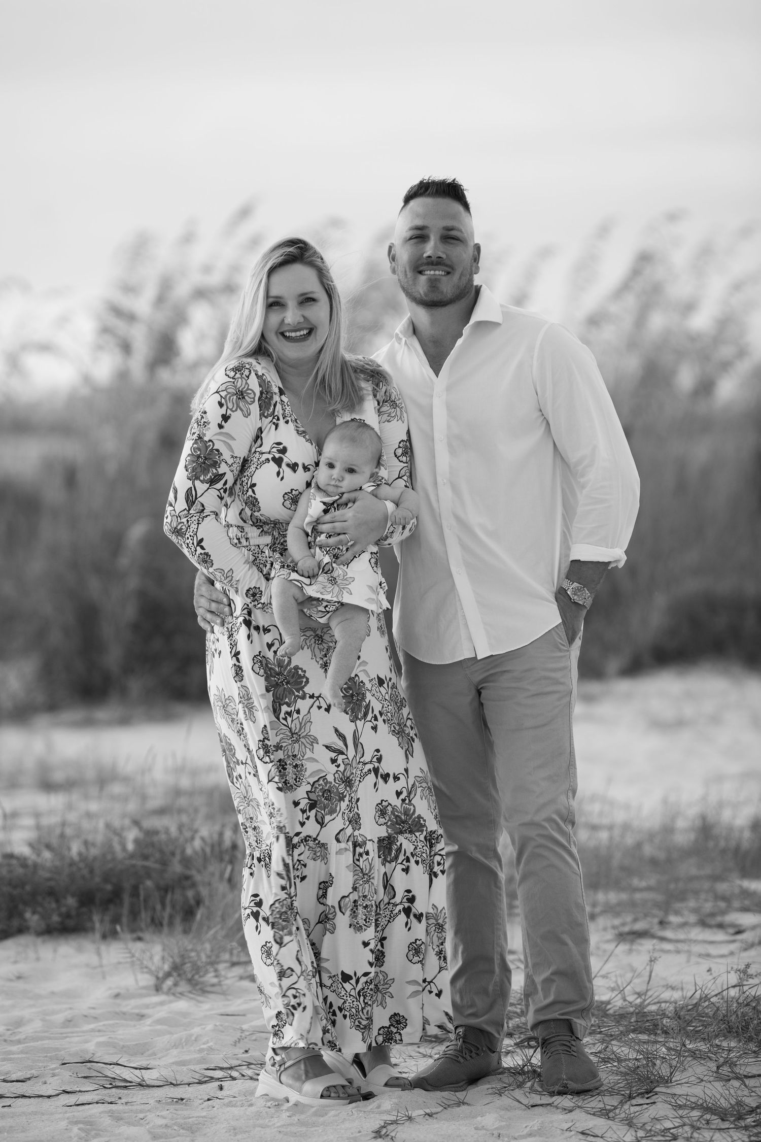 Black and white portrait of a family standing together on a sandy beach with sea oats in the background.