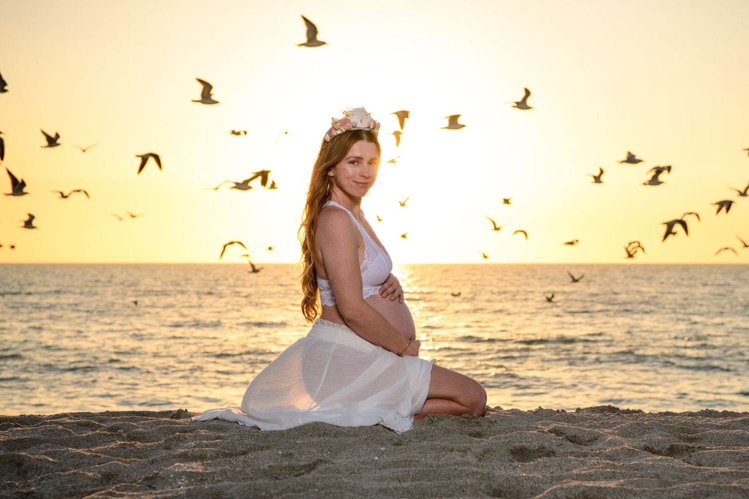 Maternity photoshoot, Woman in white dress on beach with seagulls flying at sunset.