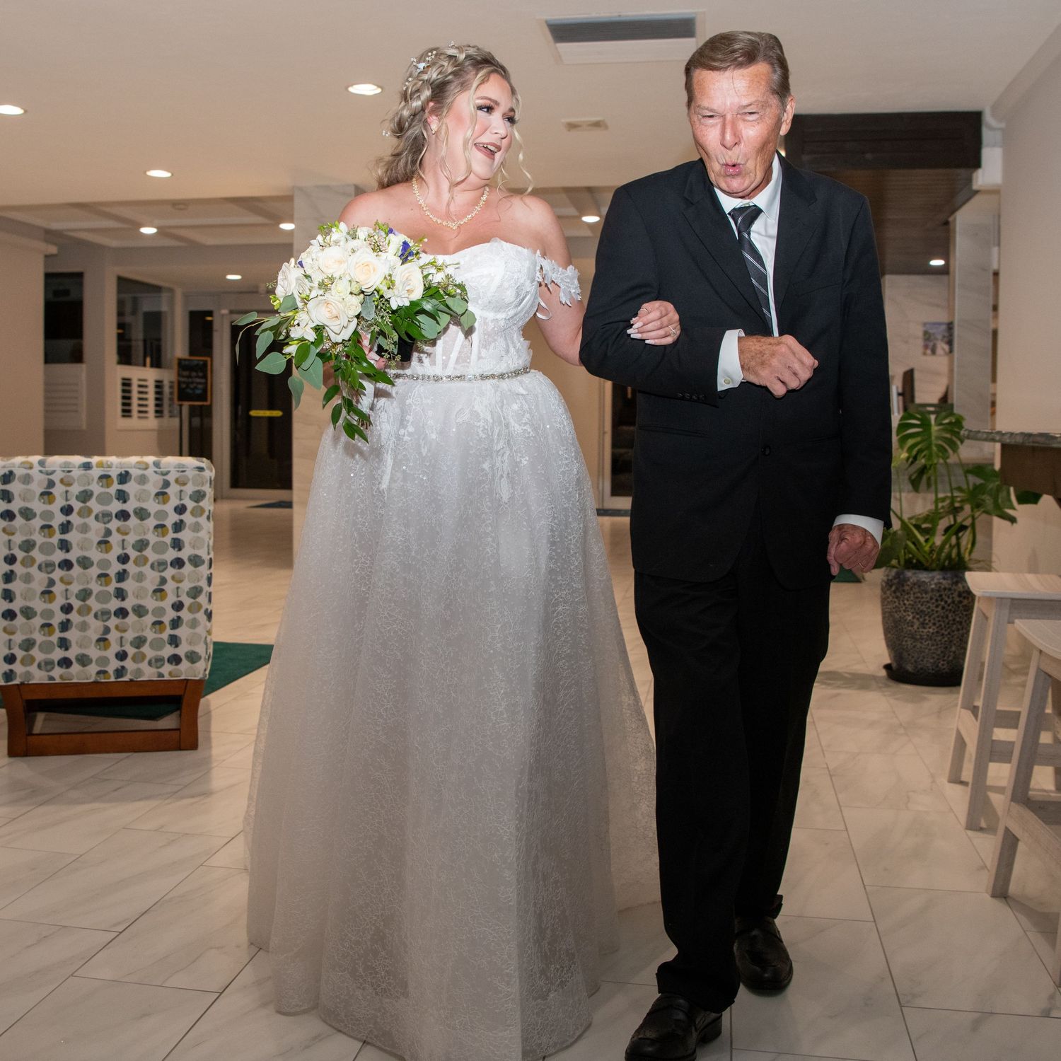 A bride in a white strapless gown walks down a bright indoor hallway with decorative wall patterns.