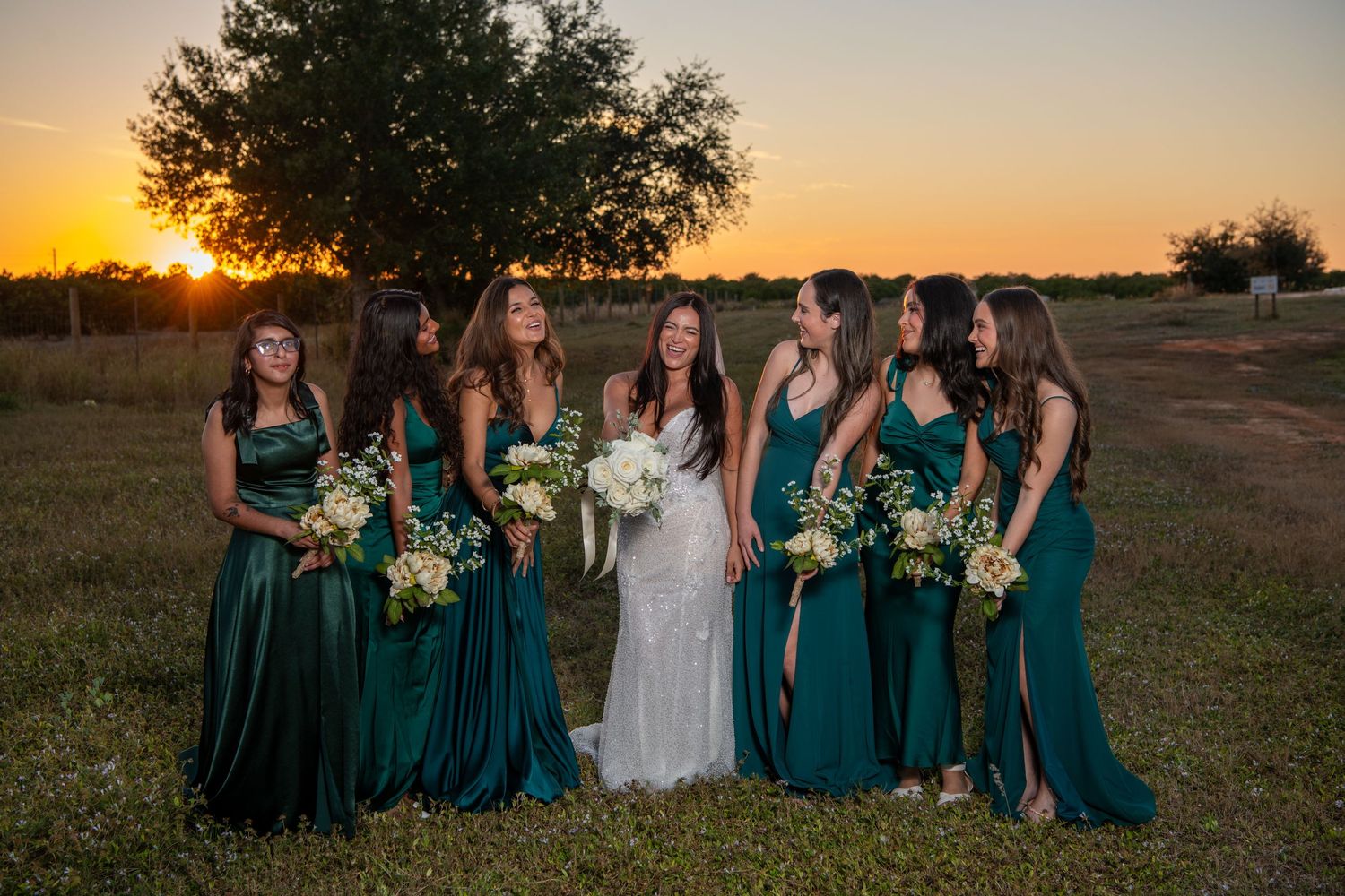 Bridesmaids in deep teal gowns gather around bride for sunset portrait session. Wedding photography.