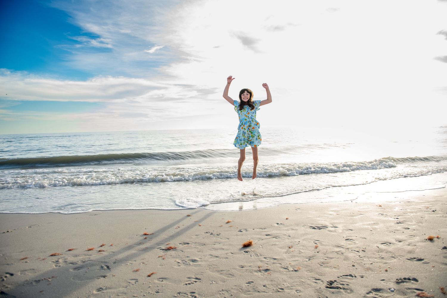 A joyful person jumps high on the beach with ocean waves and blue sky in the background.