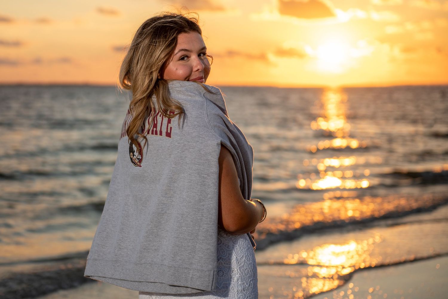 Graduate wearing FSU sweatshirt poses on beach during golden hour sunset.