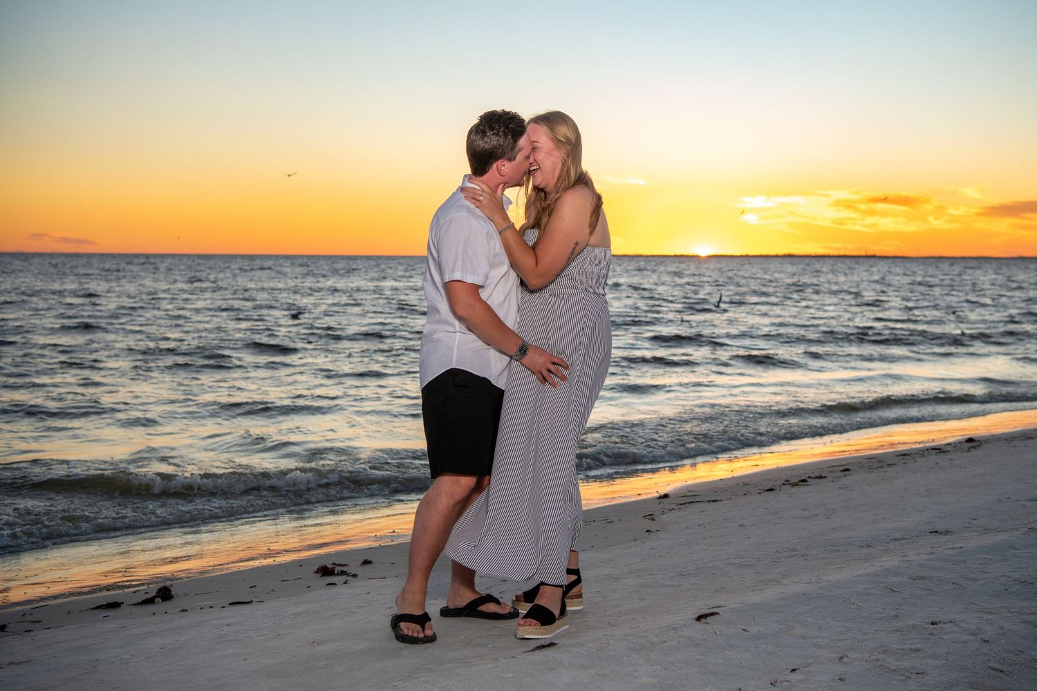 Beach engagement photoshoot with couple silhouetted against vibrant orange sunset sky.