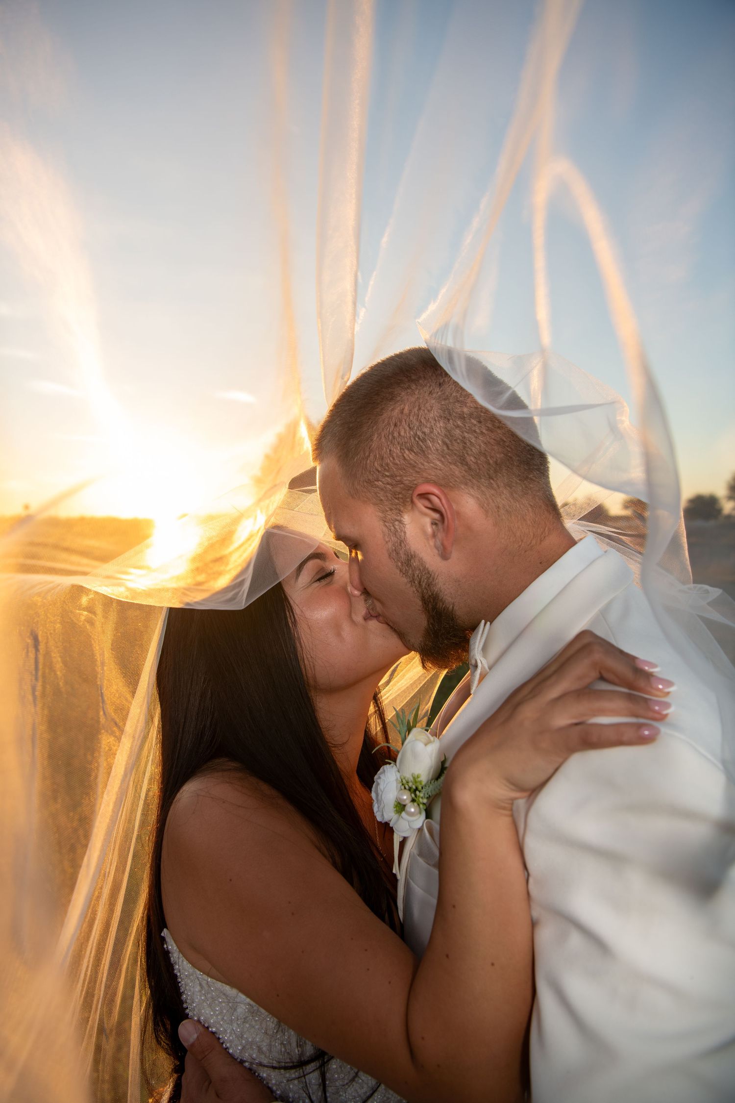 Romantic couple shares intimate kisses during golden hour sunset with veil flowing in the breeze. Wedding photography.