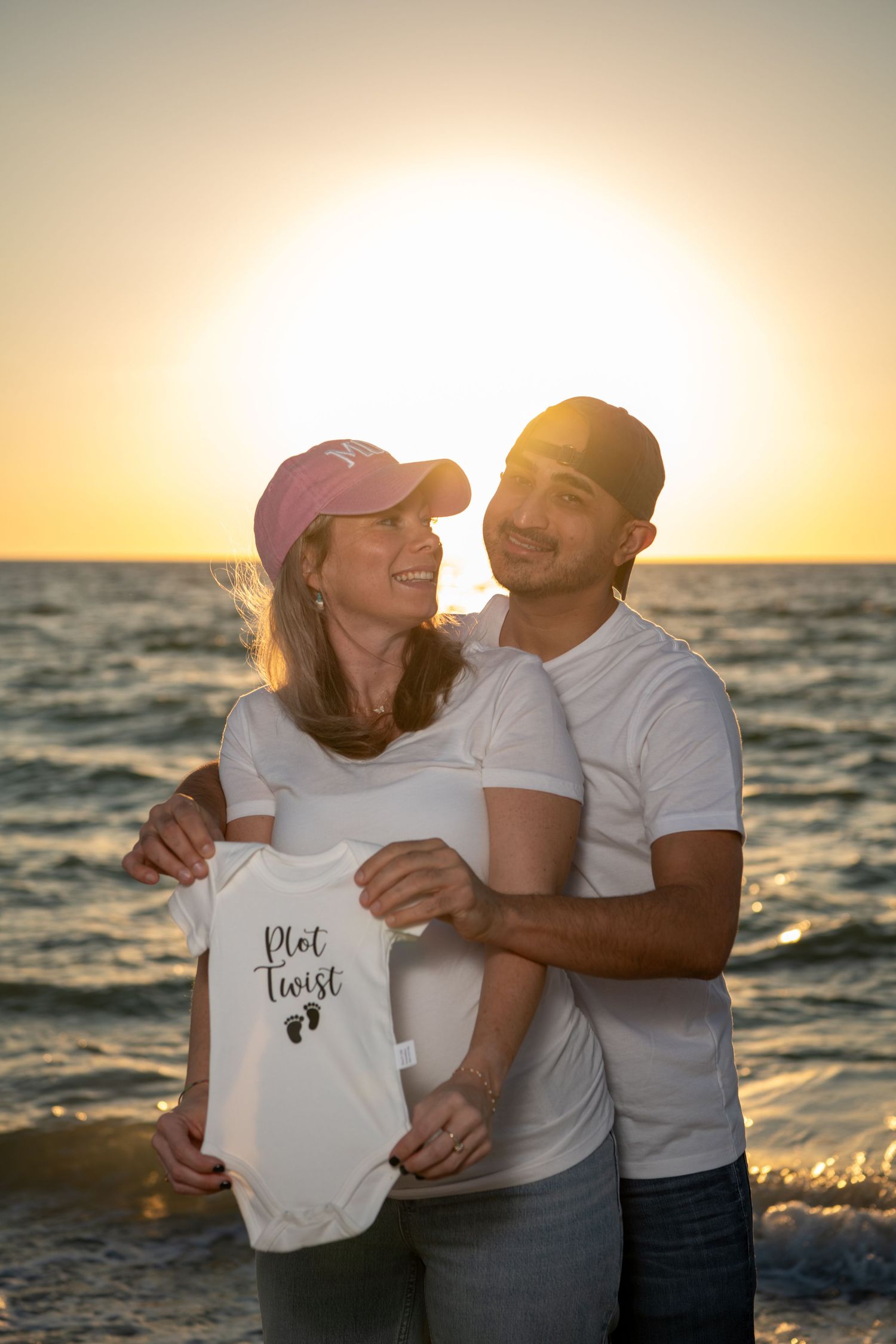 Expectant couple shares tender moment while holding baby onesie at beach. Maternity photoshoot. Couples photos.