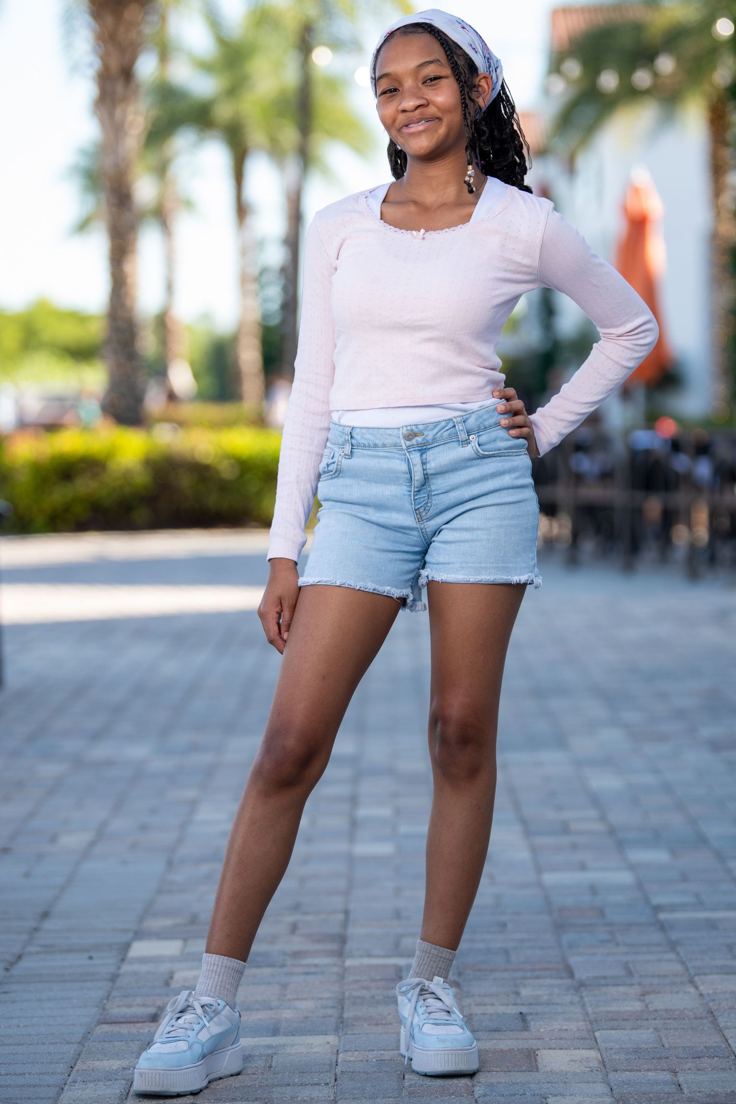 Young person in white top and denim shorts poses confidently on brick pavement. Headshots.
