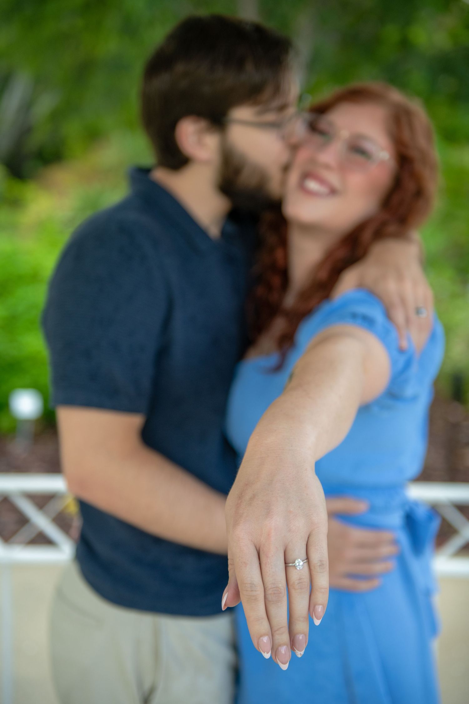 Close-up of an engagement ring being shown while couple embraces in a garden setting.
