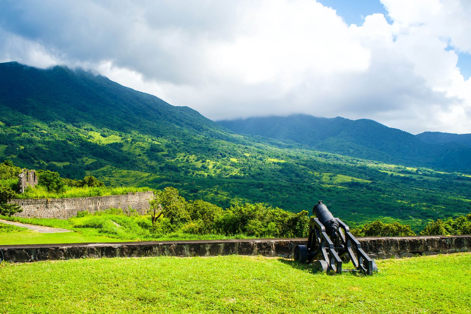Rolling green hills and mountains stretch across a scenic landscape under cloudy skies in a tropical setting.