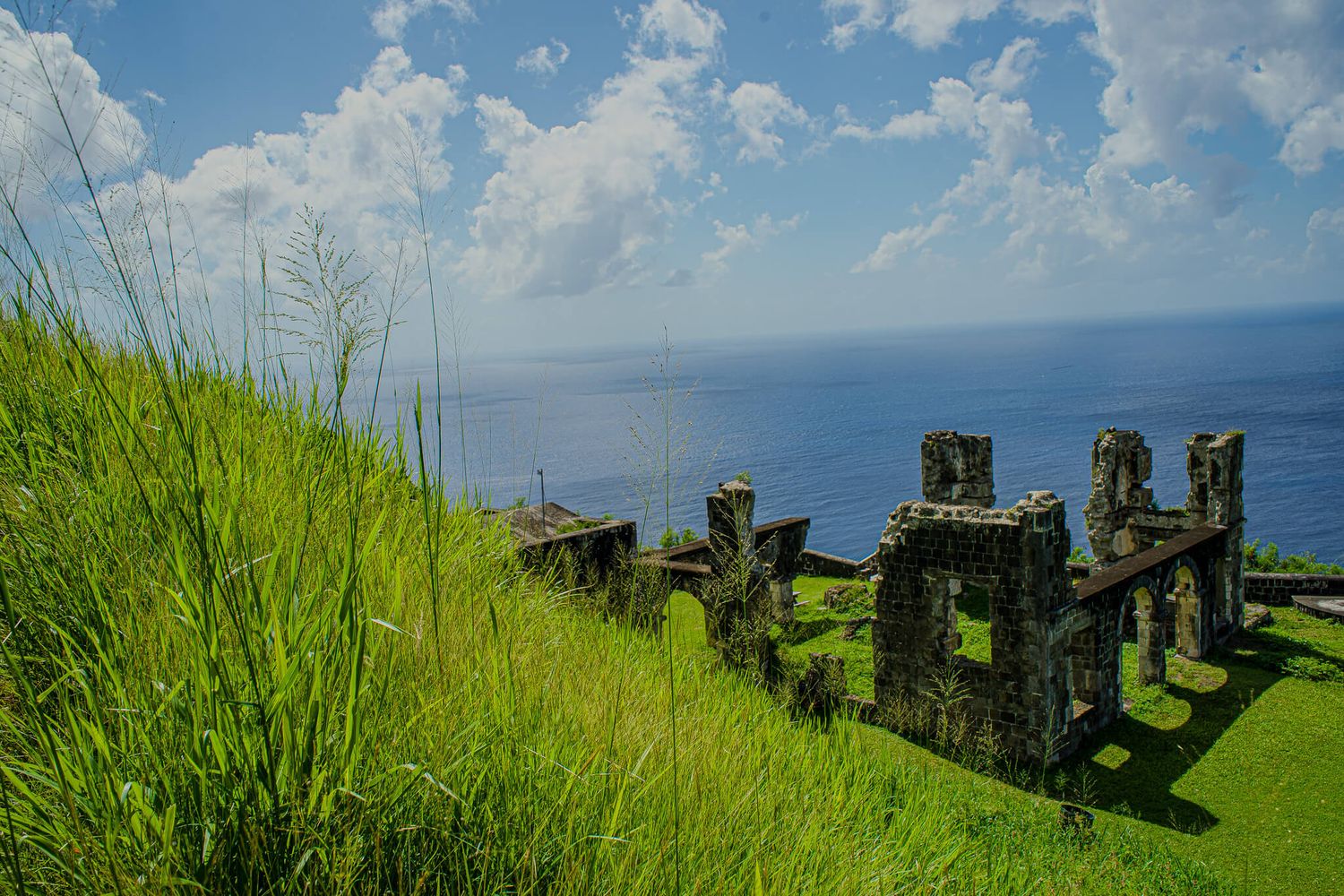 Ancient stone ruins of Brimstone Hill in St. Kitts stand atop a grassy cliff overlooking the blue ocean and sky on a sunny day.