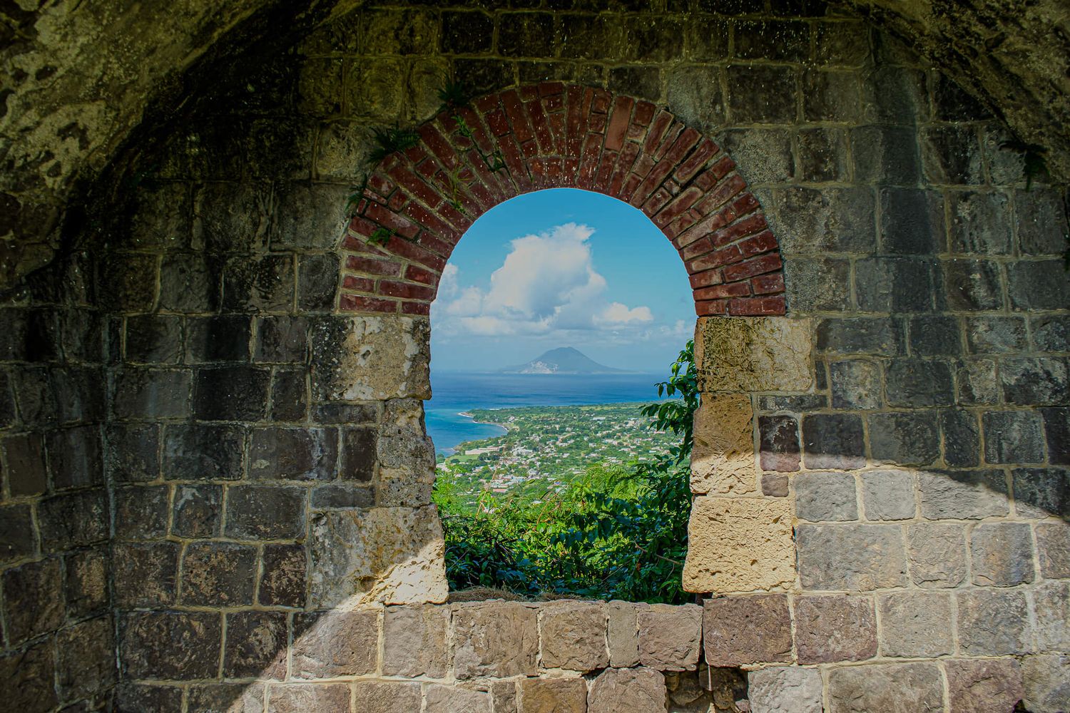 Stone archways of an ancient fortress overlook a bright blue Caribbean sea and lush tropical landscape.