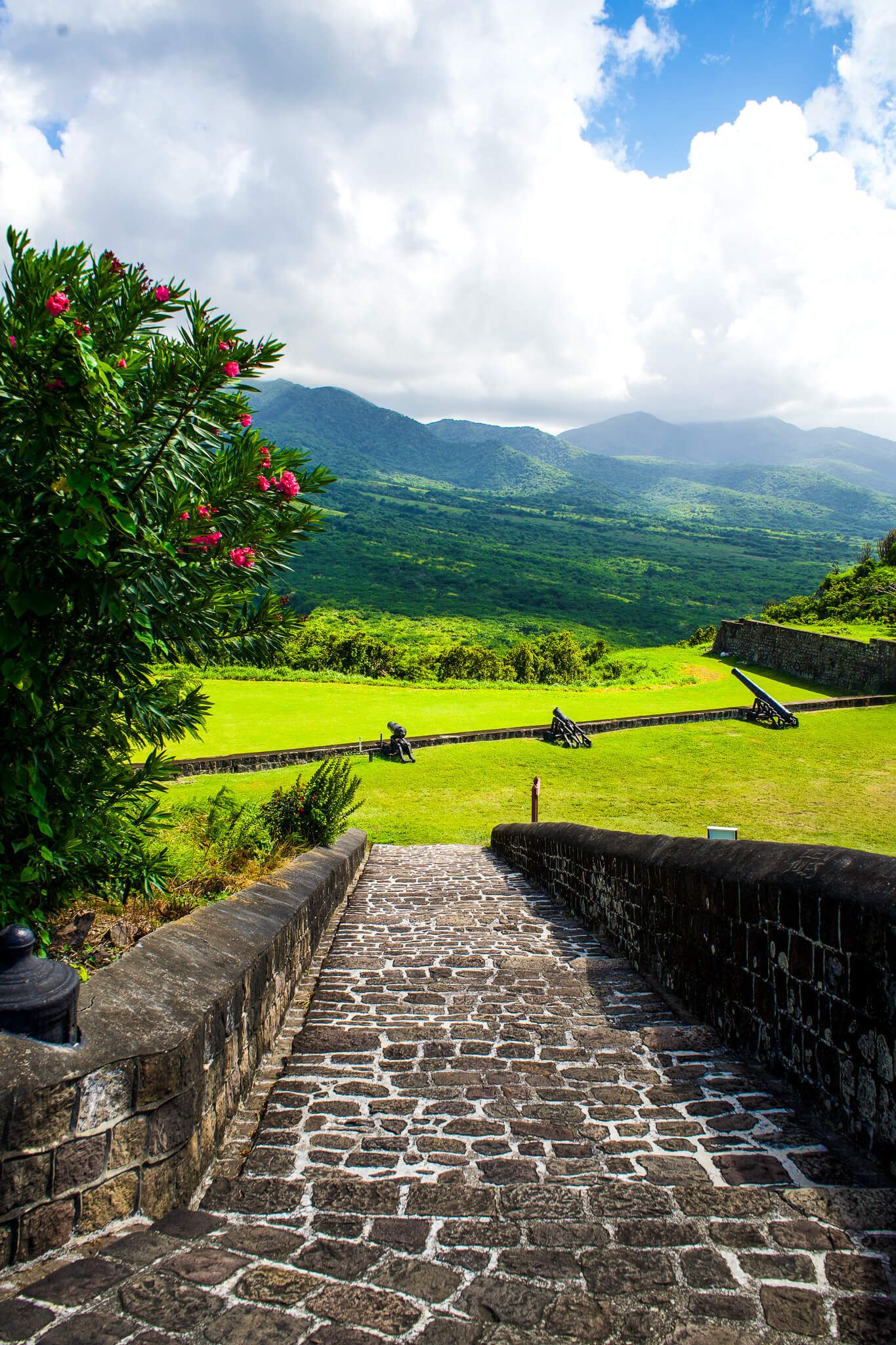 Stone pathway along fortress walls overlooking lush green valley and mountains in Saint Kitts under cloudy skies.