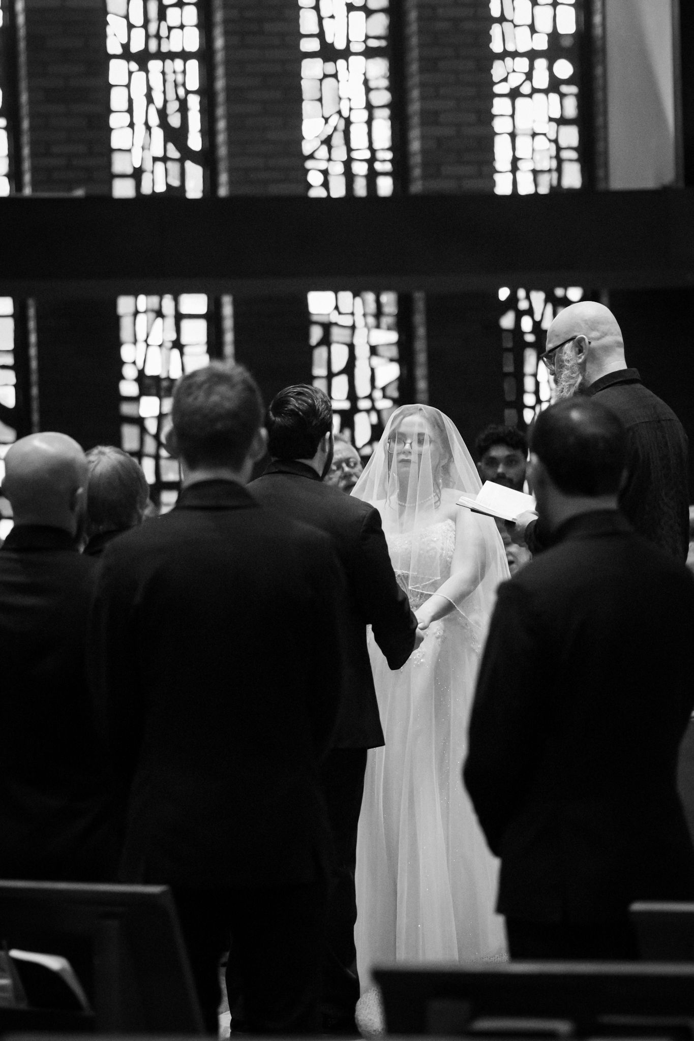 A black and white photo shows wedding attendees in formal attire standing in a church with ornate stained glass windows.
