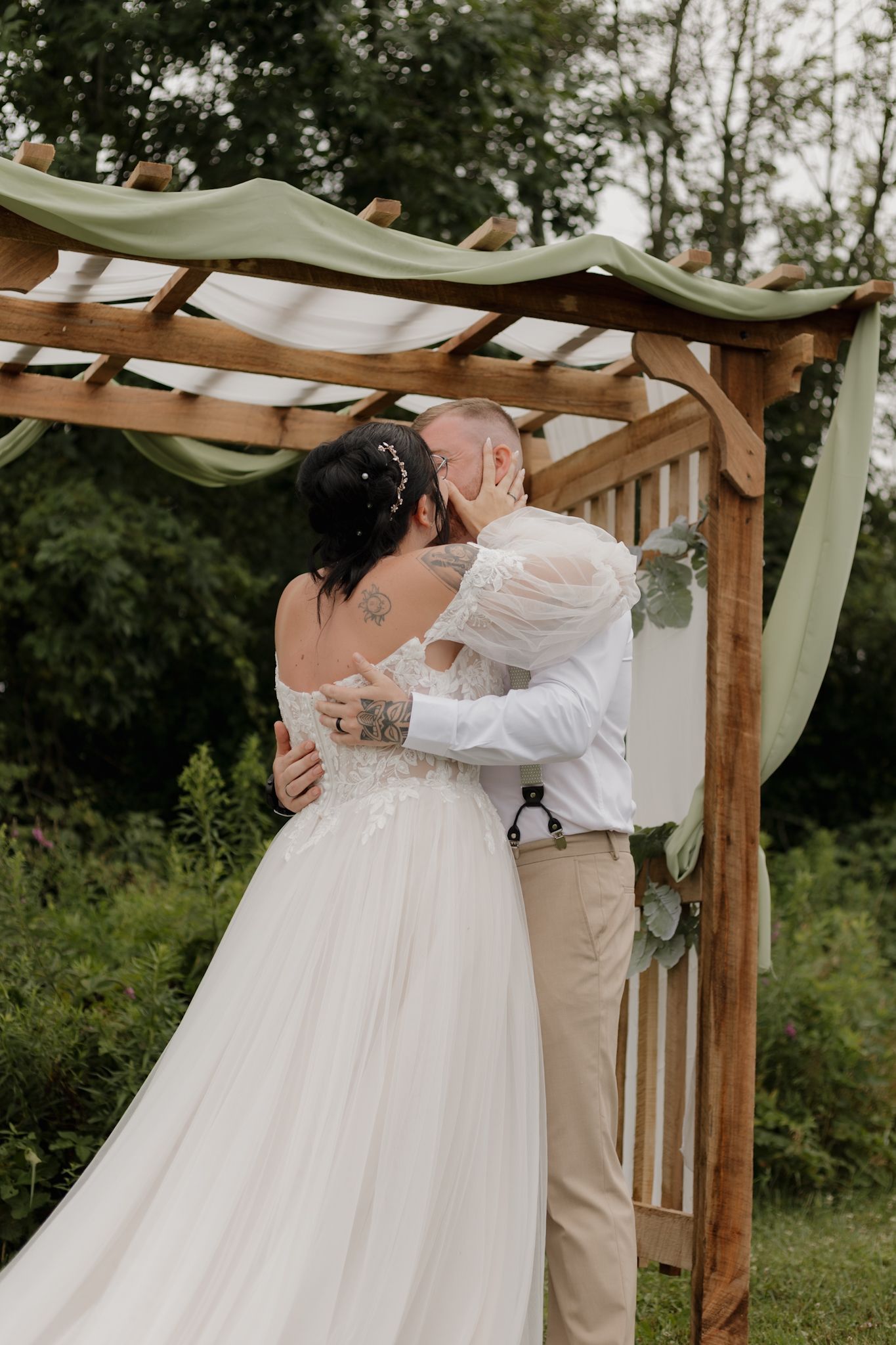 A romantic wedding ceremony under a wooden pergola draped with sage green fabric in an outdoor garden setting.