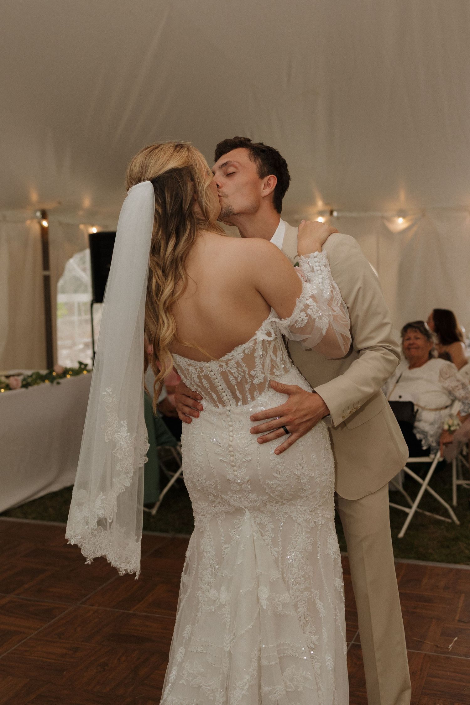 Bride and groom sharing first dance kiss under tent at wedding reception, bride in lace backless gown with veil.