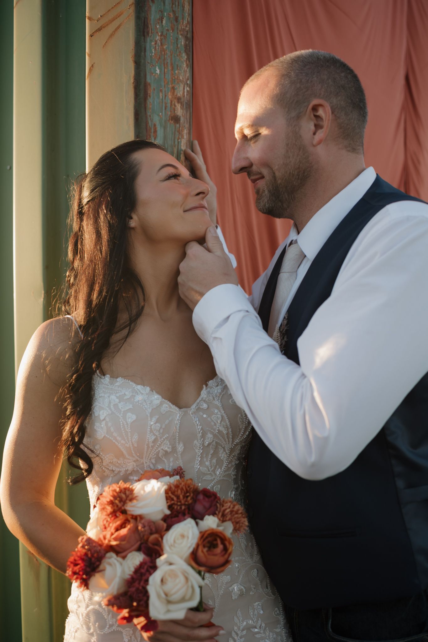 A groom tenderly holds his bride's chin as she smiles up at him, bride holding a bouquet of red and white roses.