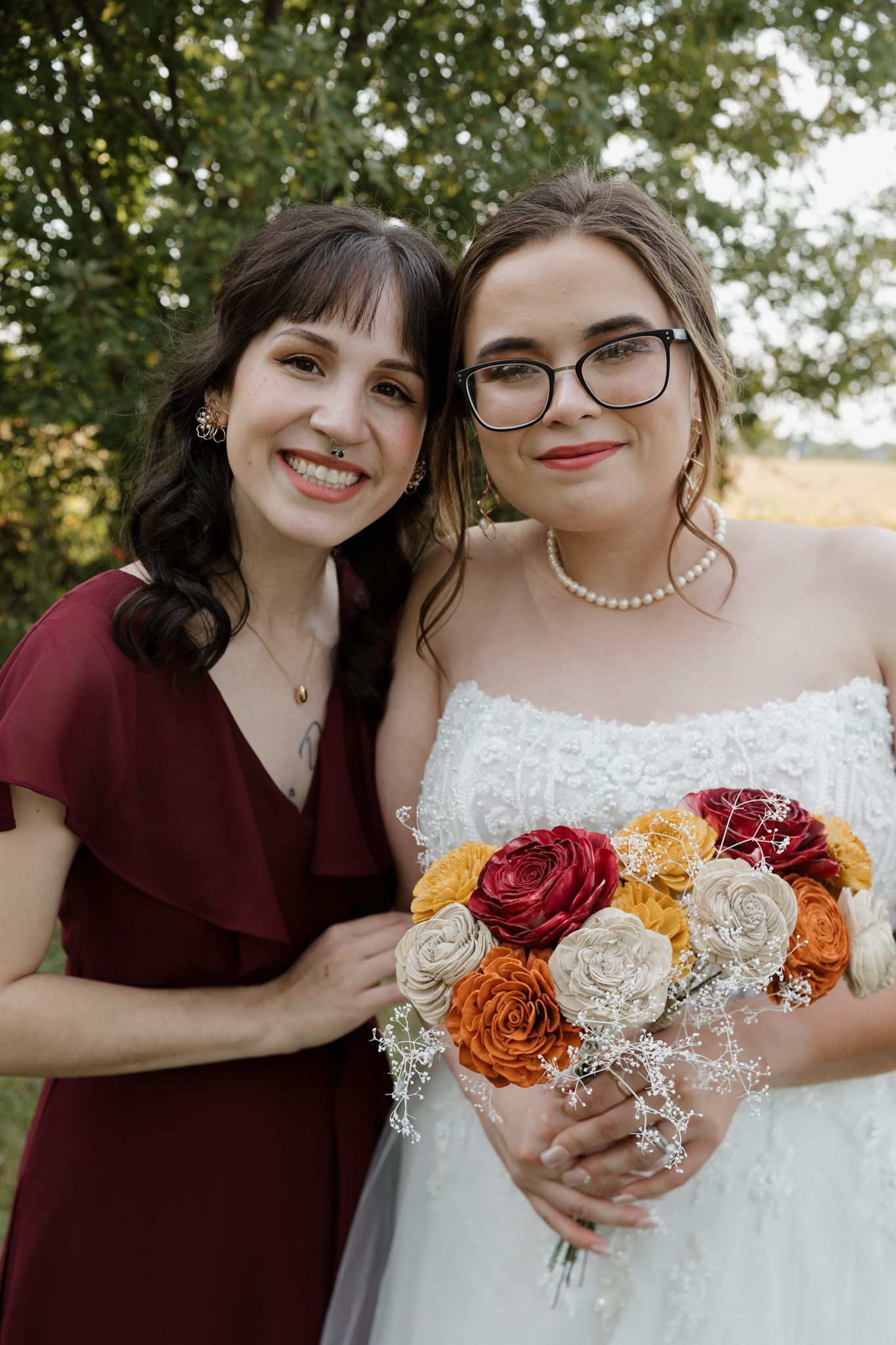 A bridal bouquet featuring red, orange and white roses held between two people in burgundy and white attire outdoors.
