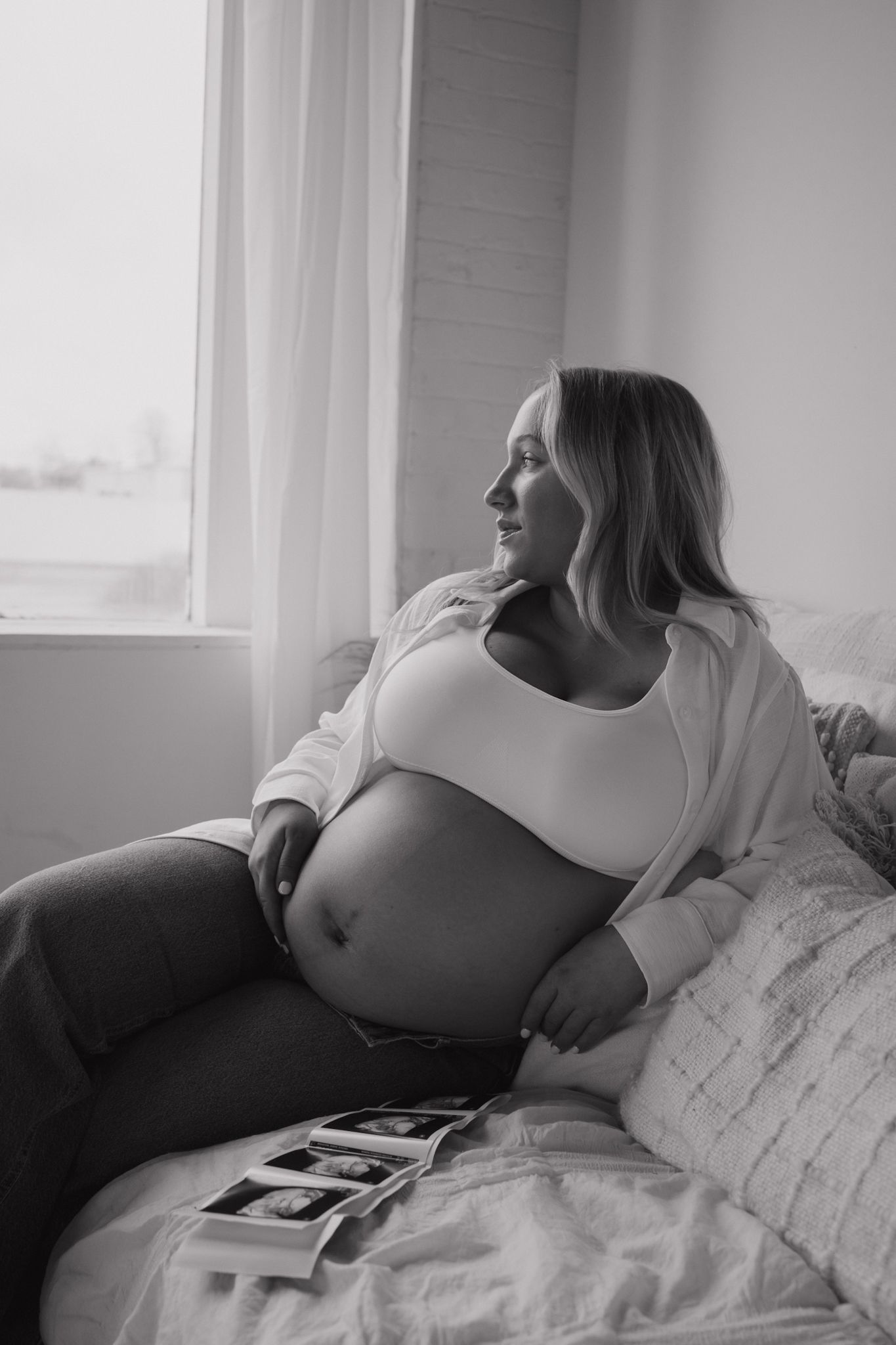 Black and white photo of a pregnant woman sitting on a bed, holding her baby bump while gazing out a window with ultrasound photos.