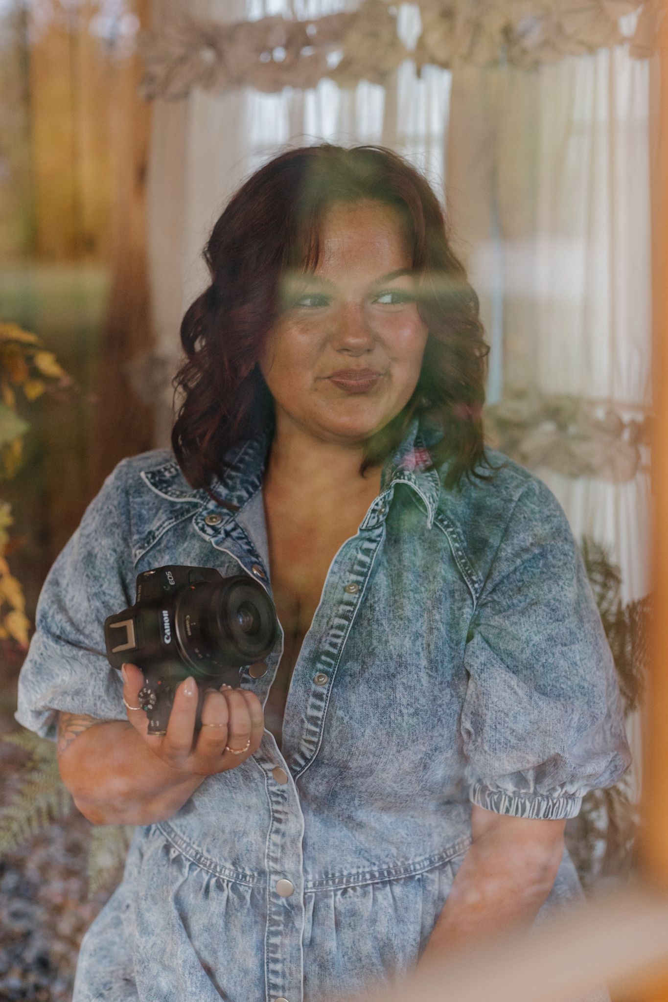 A photographer smiles while holding a camera in a denim dress with sunlit curtains and flowers in the background.