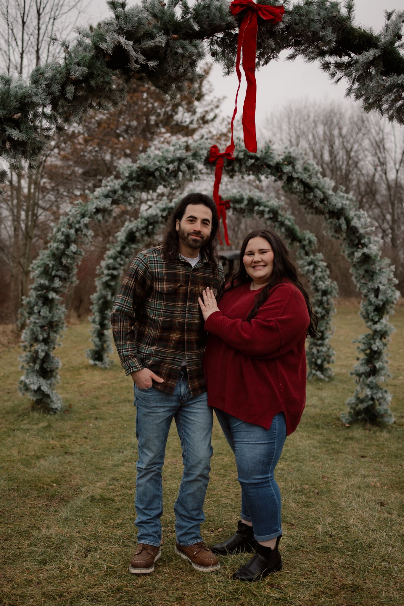 A couple in casual winter attire poses together under a festive evergreen arch decorated with a red bow outdoors.