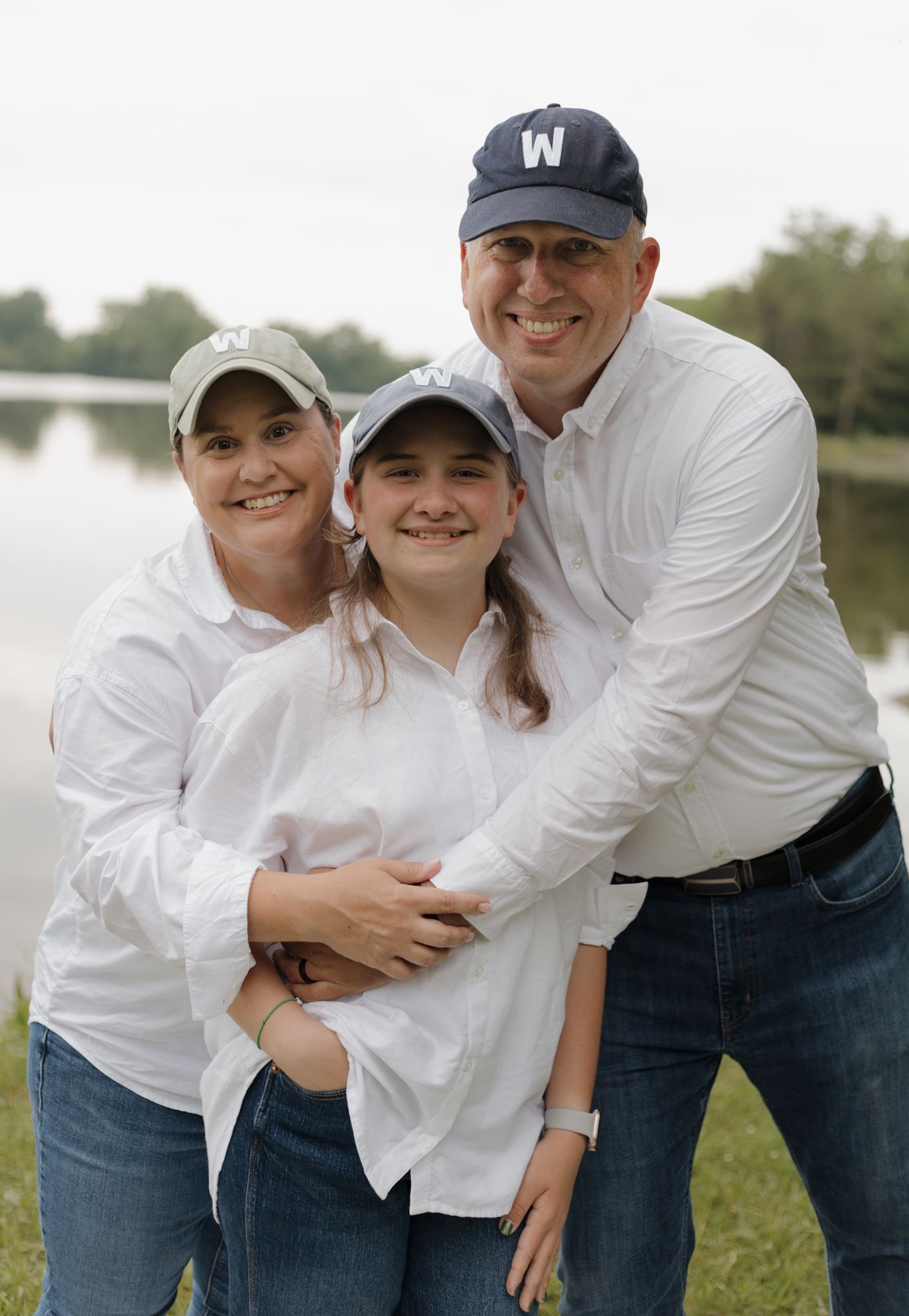 Family of three wearing matching white shirts pose together by a lake on a sunny day.