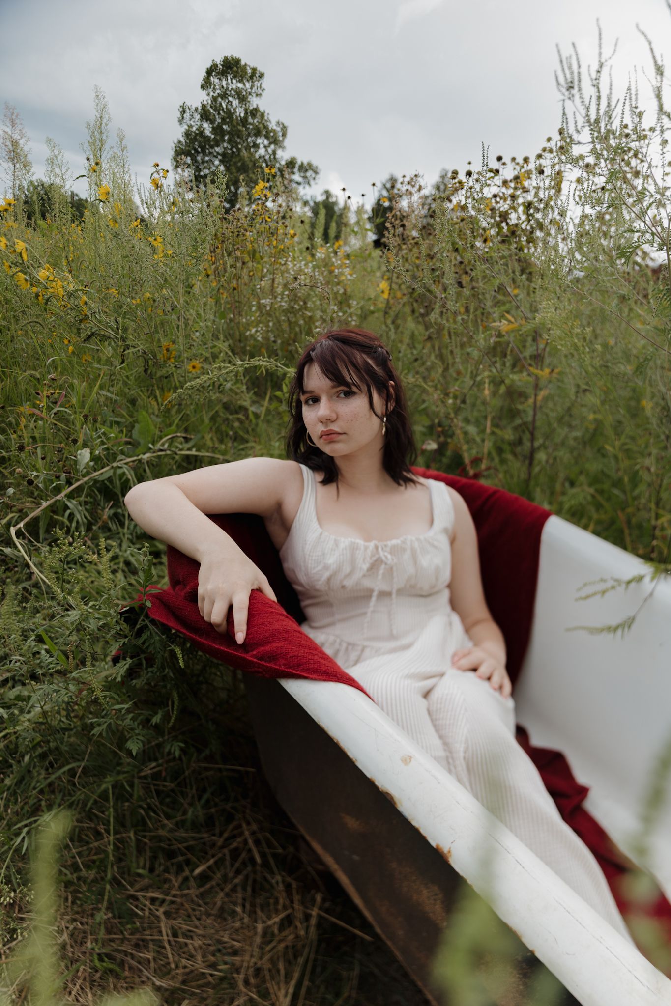 A person in a white dress reclines on a burgundy velvet couch outdoors among tall wildflowers and natural greenery.