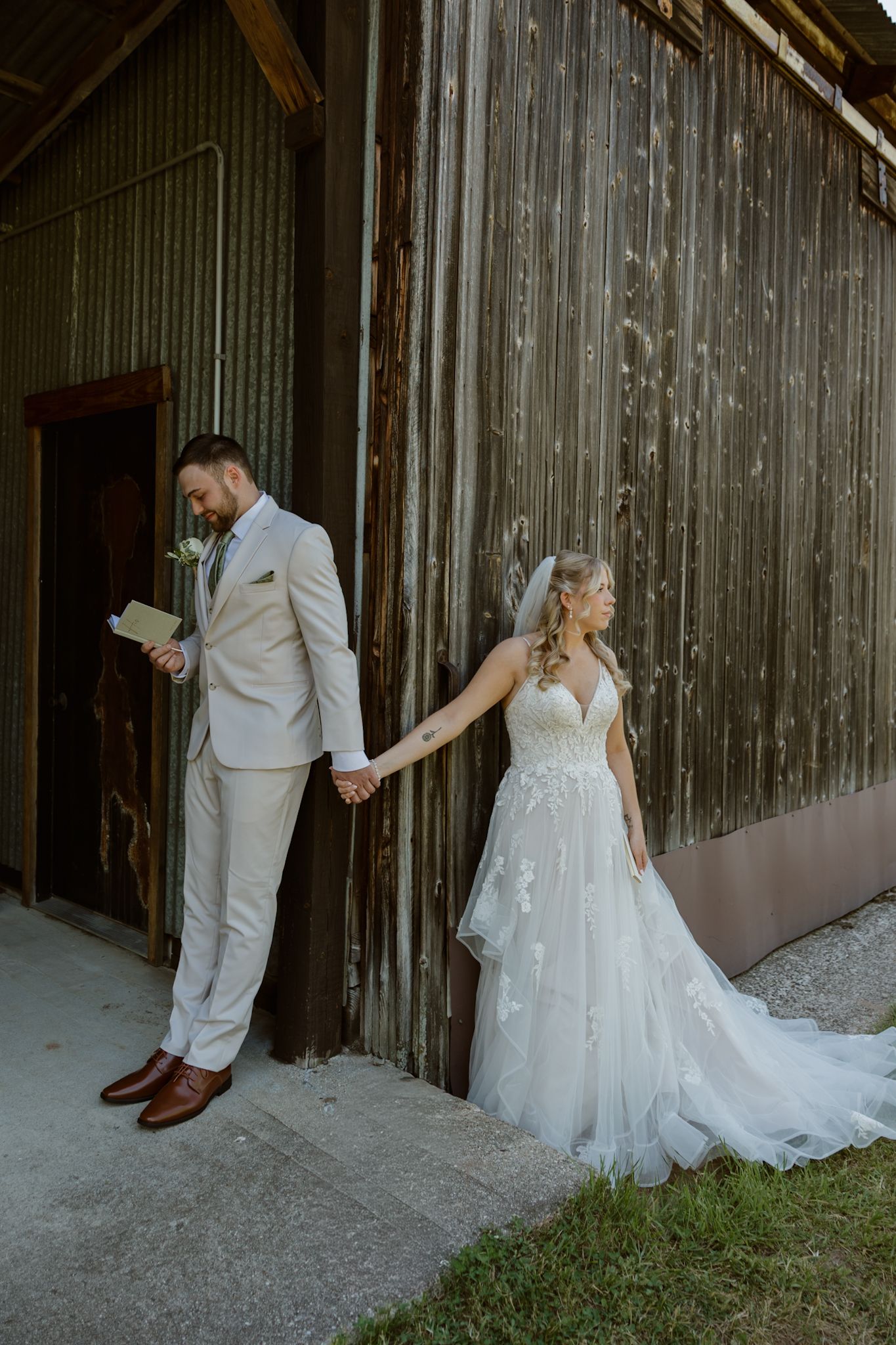 Bride in white lace gown and groom in tan suit hold hands outside rustic barn during wedding first look.