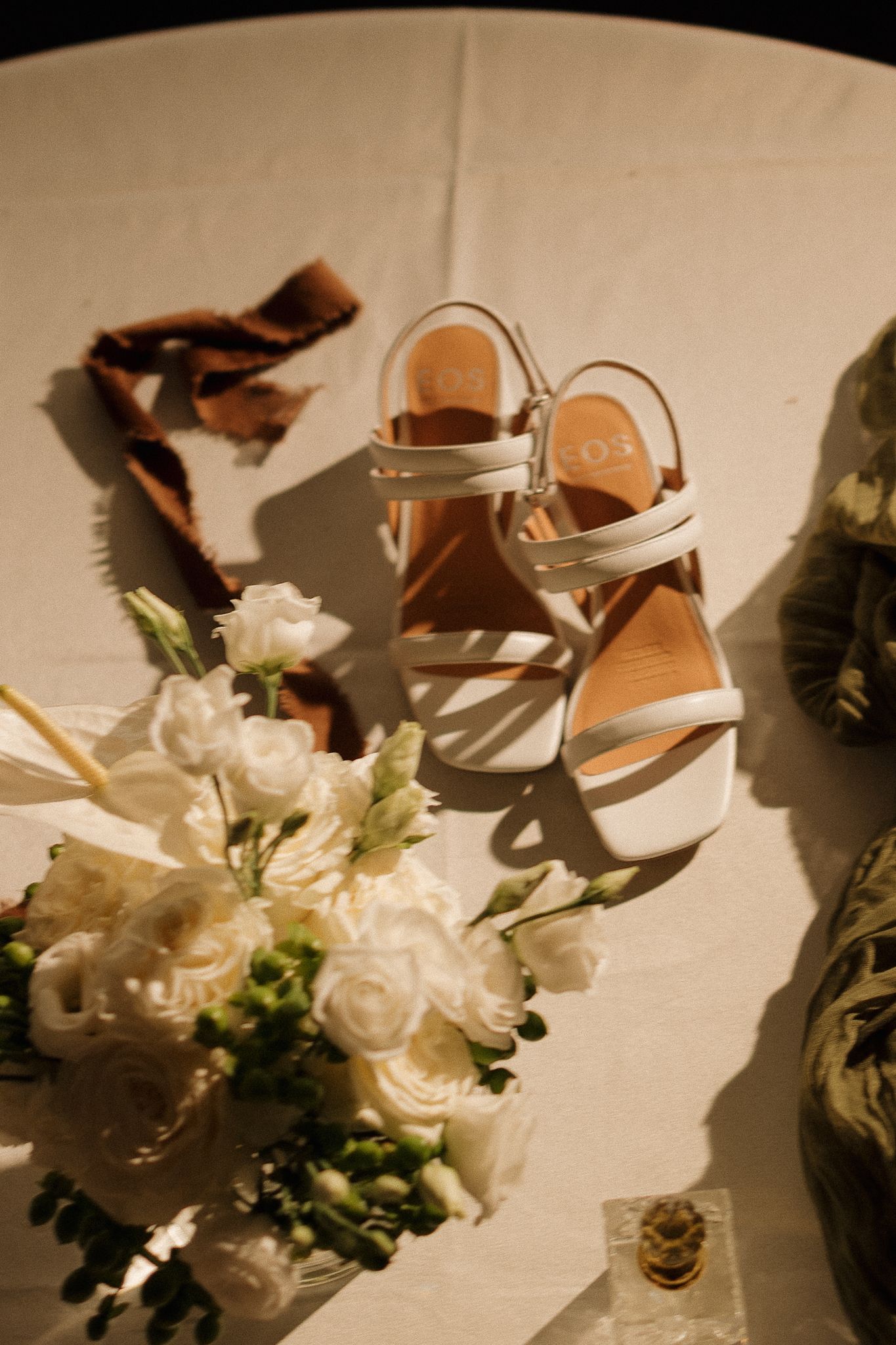 White bridal sandals with tan insoles beside a white rose bouquet on a draped table.
