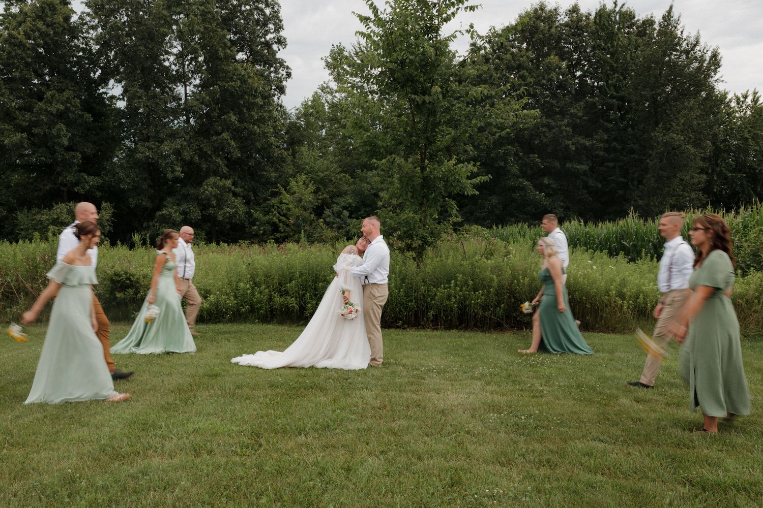 Wedding party in mint green dresses poses on a grassy lawn surrounded by trees during an outdoor ceremony.