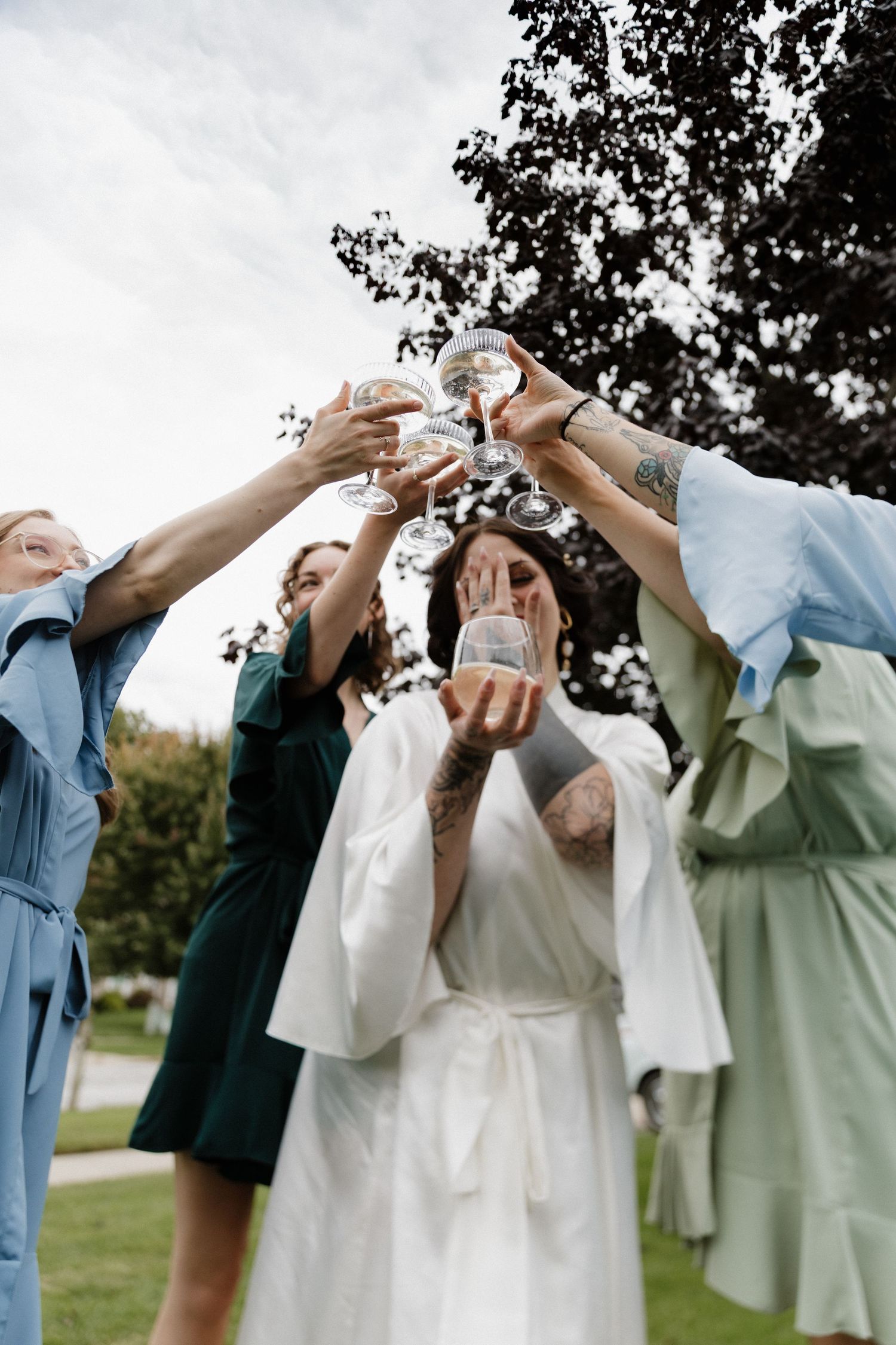Wedding party members in light colored dresses clink champagne glasses together in celebration outdoors.