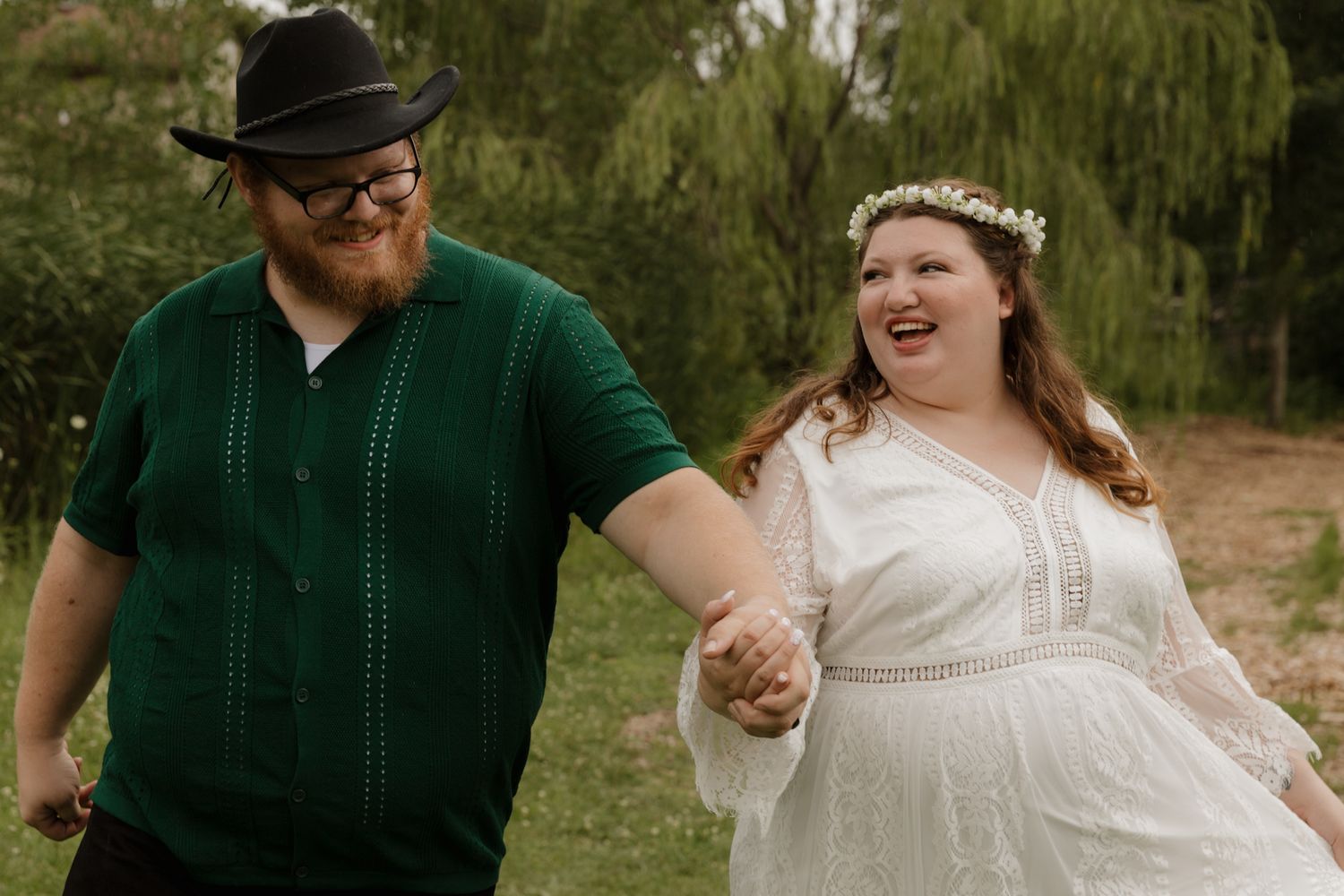 A couple dances joyfully outdoors wearing wedding attire in a natural setting.