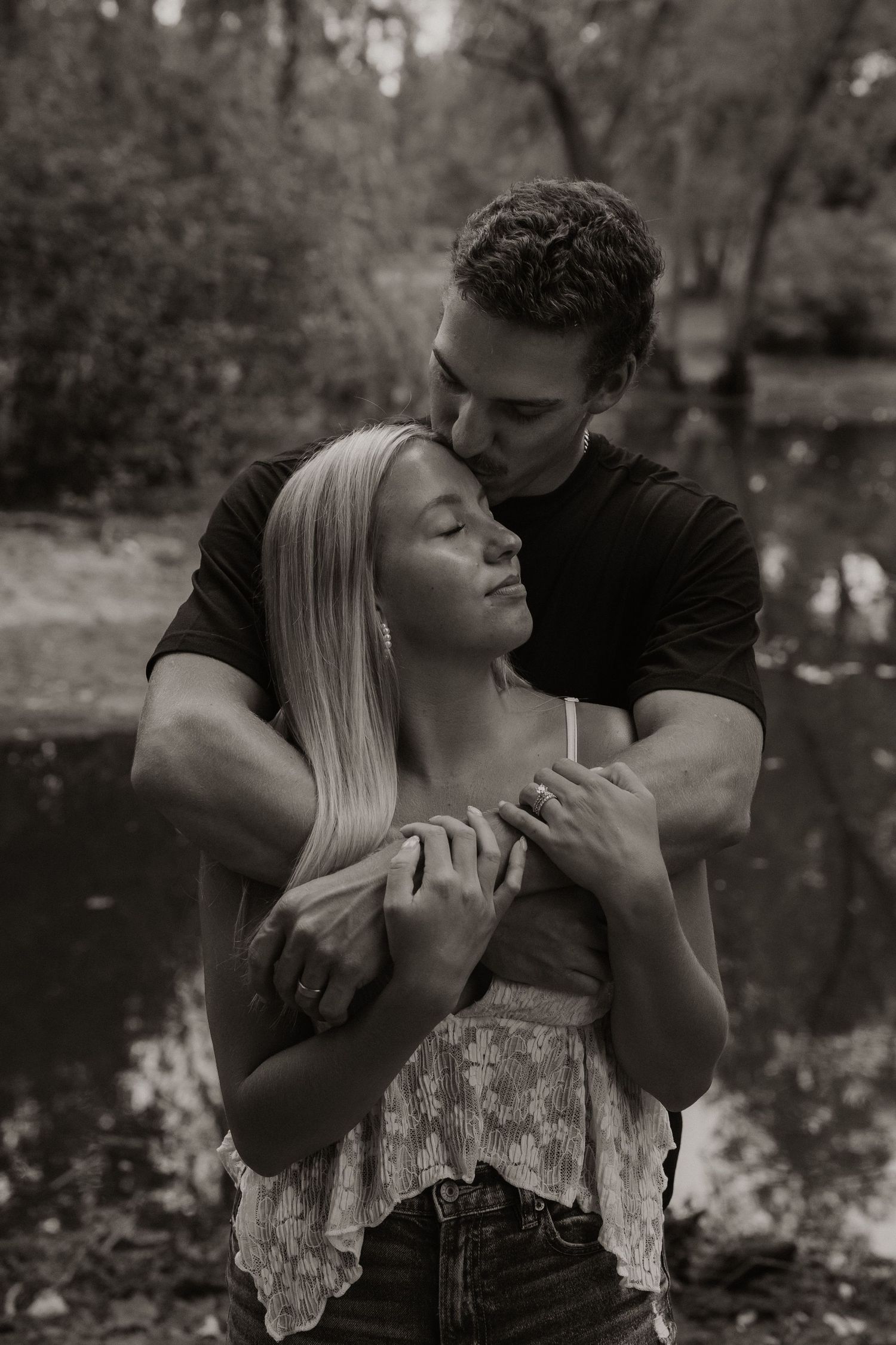 Black and white photo of a couple embracing outdoors, man kissing woman's temple near a serene forest pond.