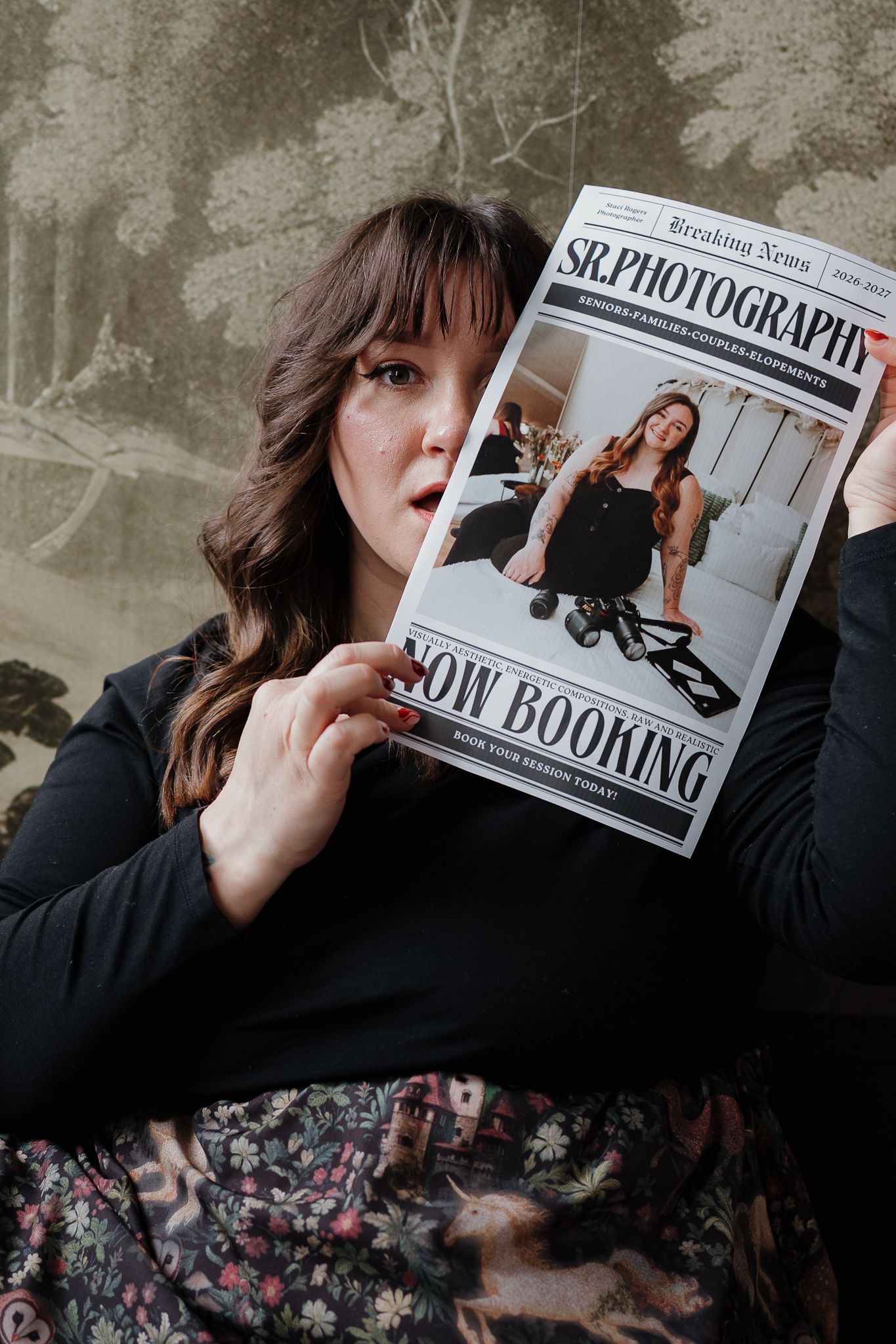 Woman holding SR Photography newspaper mock-up showing Now Booking announcement against decorative background.