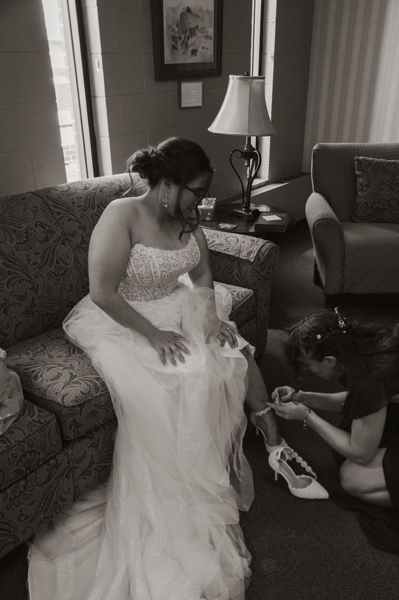 Black and white photo of a bride in a strapless wedding gown sitting on couch while getting shoes put on.