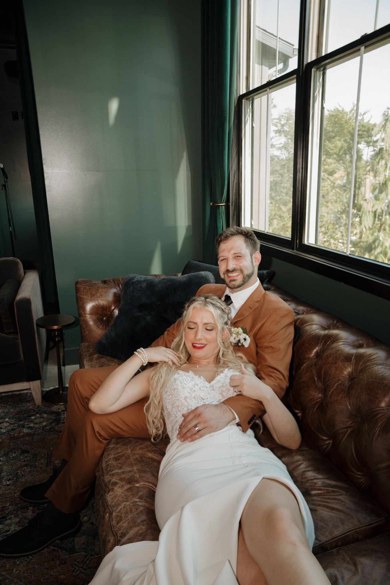 A couple shares intimate laughter while relaxing together on a brown leather couch near sunlit windows.