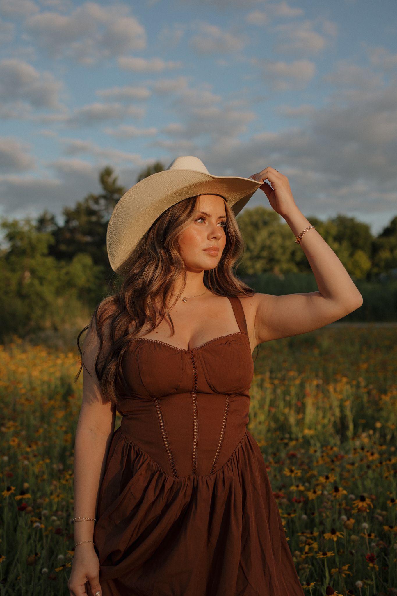 Woman in brown dress and wide-brim hat standing in a wildflower field at golden hour sunset.