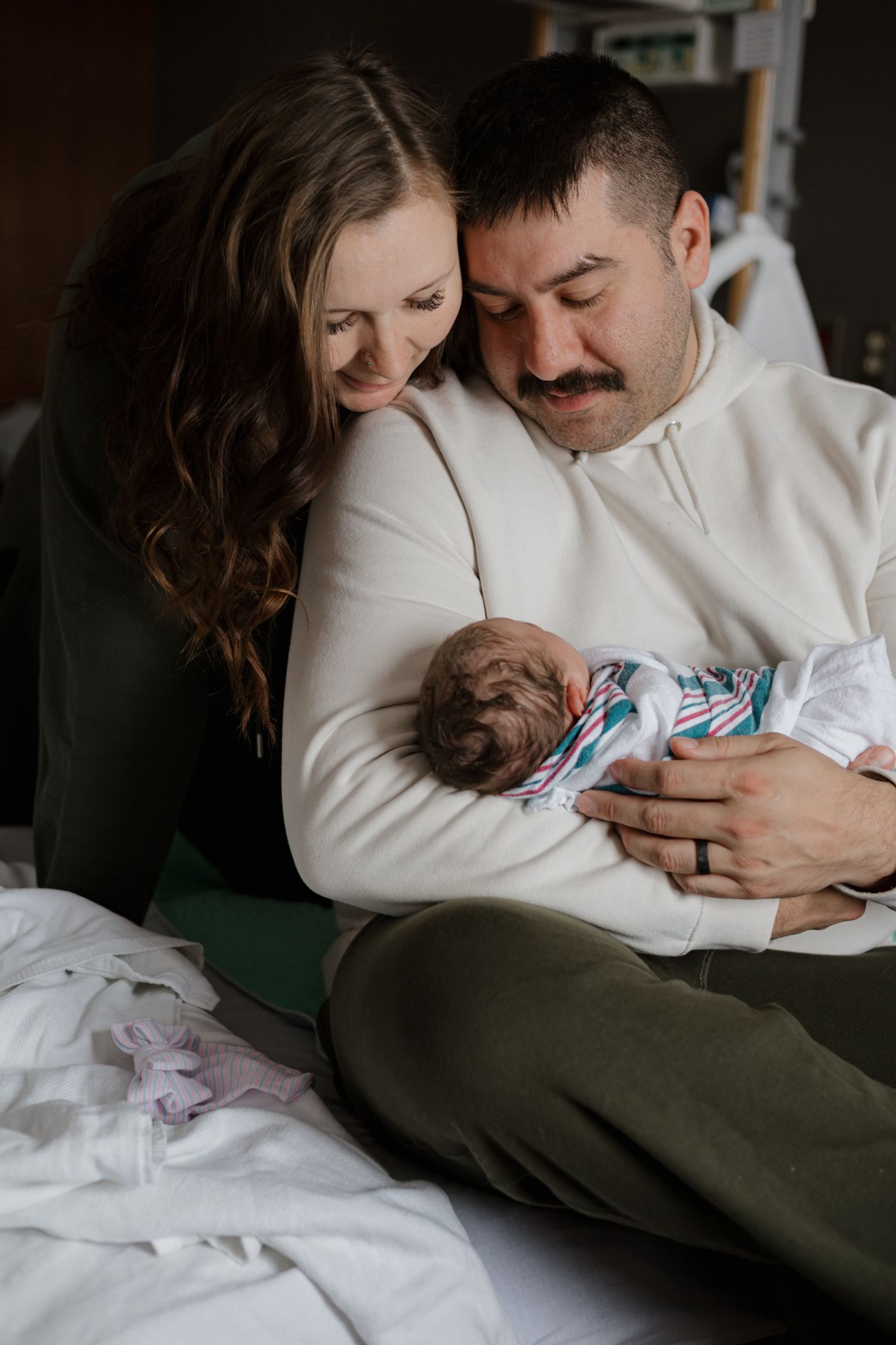 A couple in white sweaters cuddles with their newborn baby wrapped in a striped blanket while sitting on a bed.