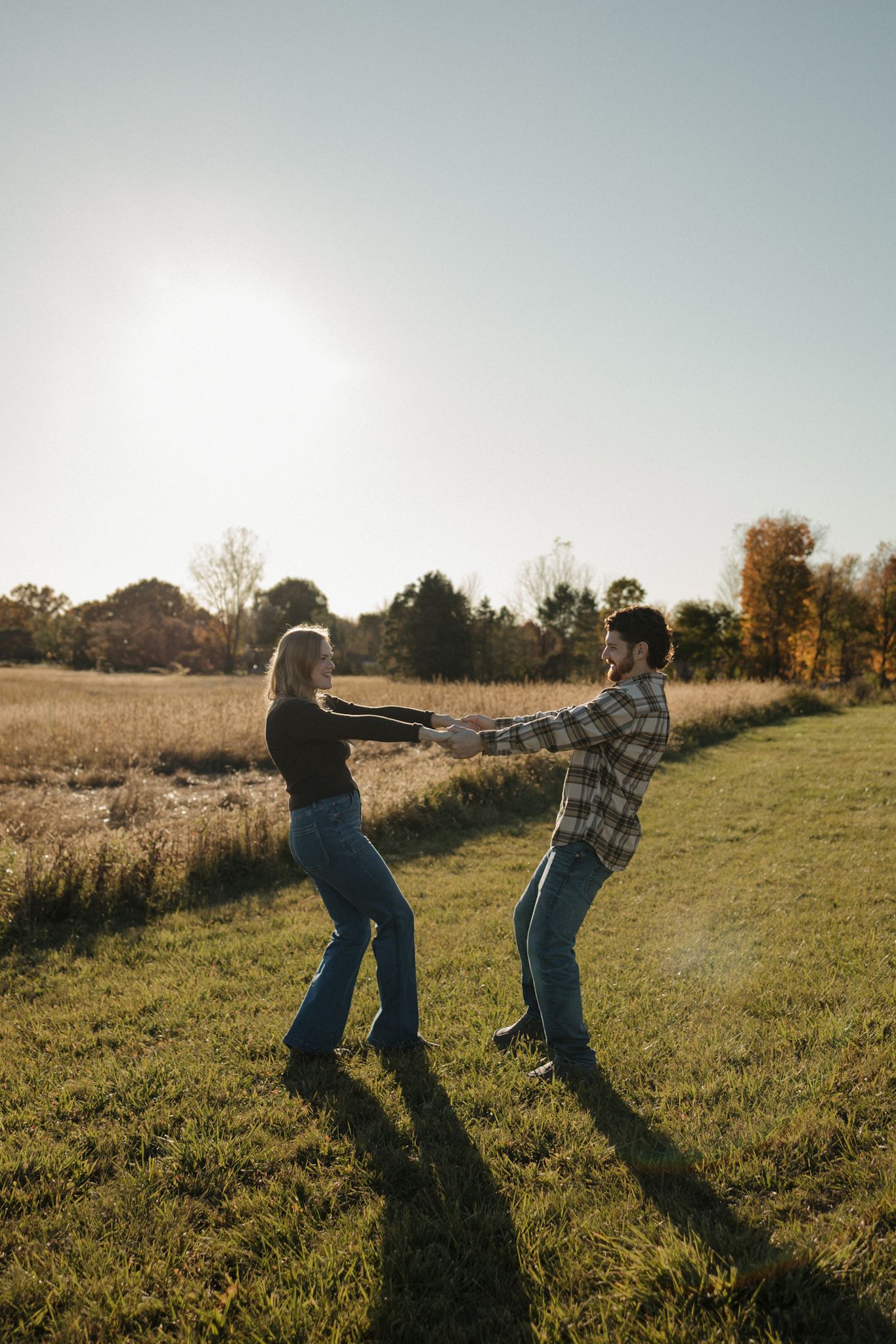 Two people playfully dance and spin together in a sunlit autumn field casting long shadows on the grass.