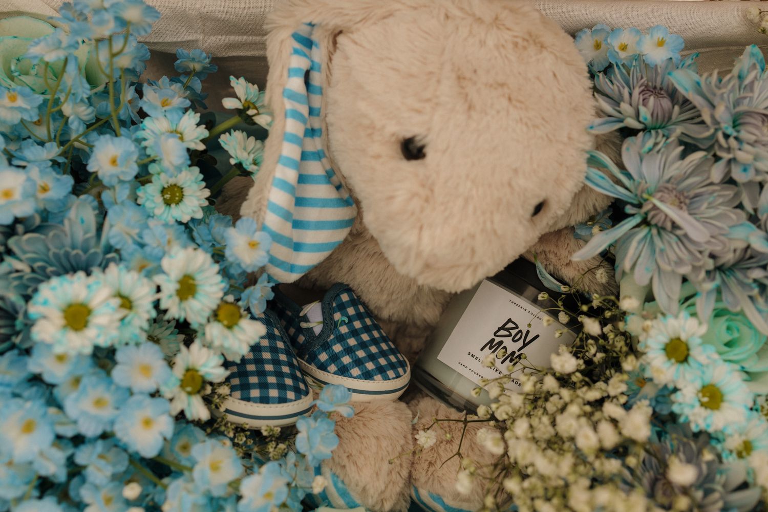 Teddy bear with striped hat surrounded by blue daisies, gingham baby shoes, and a Boy Mom candle for a baby shower.