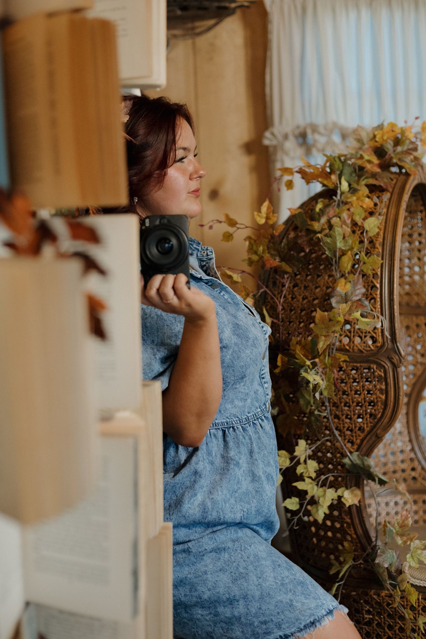 A photographer in a blue denim dress holds a vintage camera while posing near decorative floral elements.