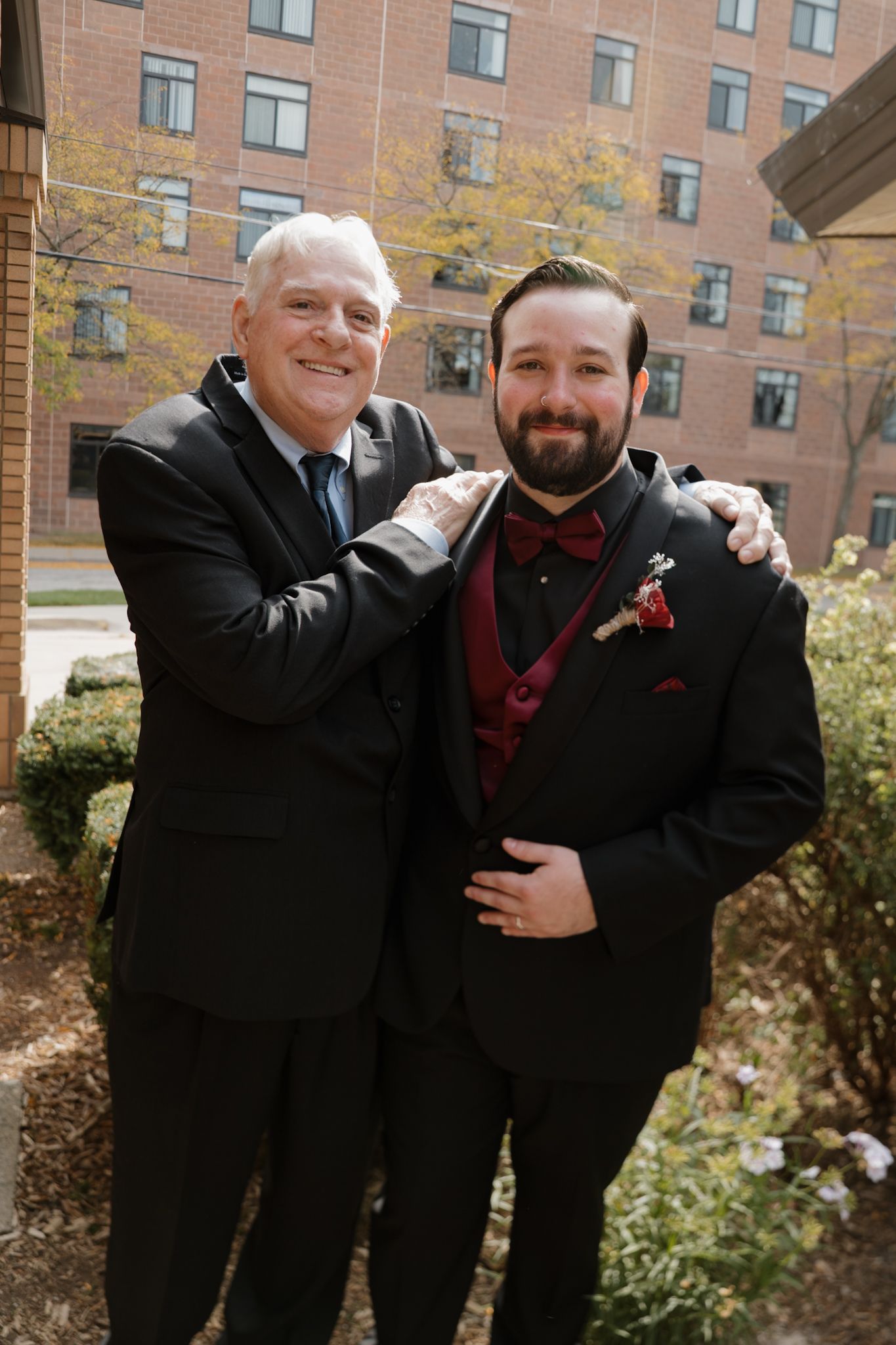 Groomsmen in black suits and burgundy accessories pose together outside a brick building.