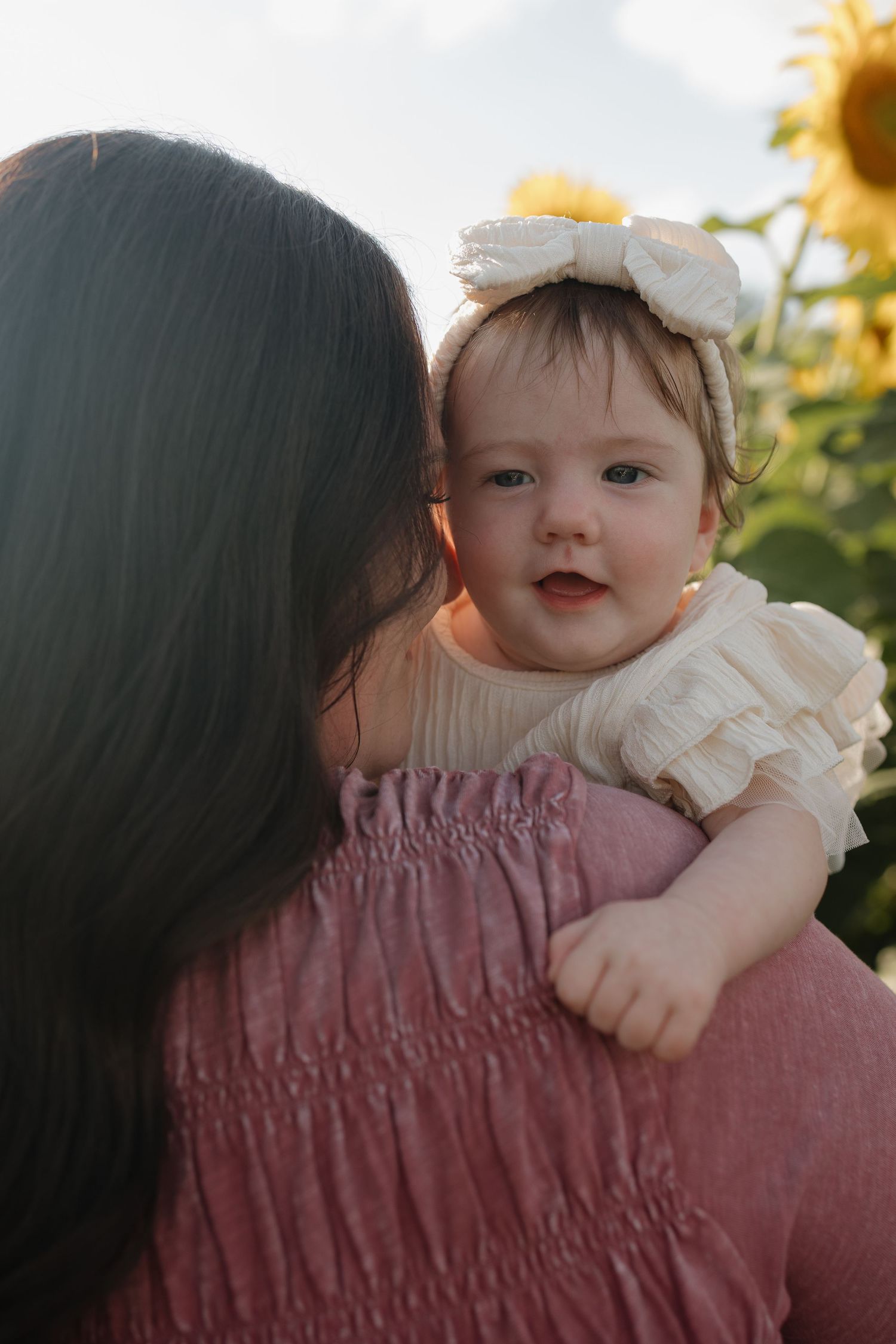 A mother holds her baby girl wearing a white bow headband in a sunflower field during golden hour.