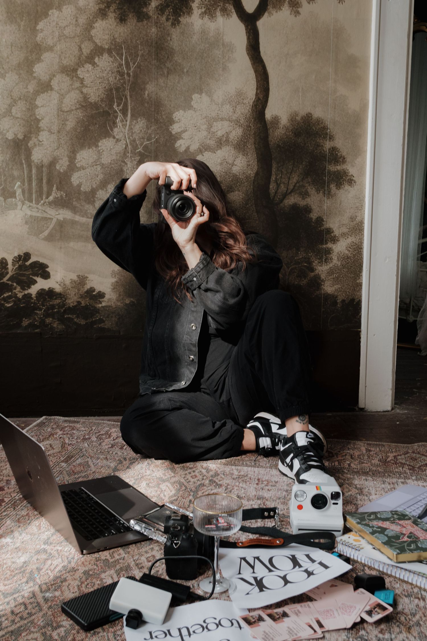Woman in black outfit sits on floor photographing with DSLR camera, surrounded by laptop, Polaroid, and magazines.