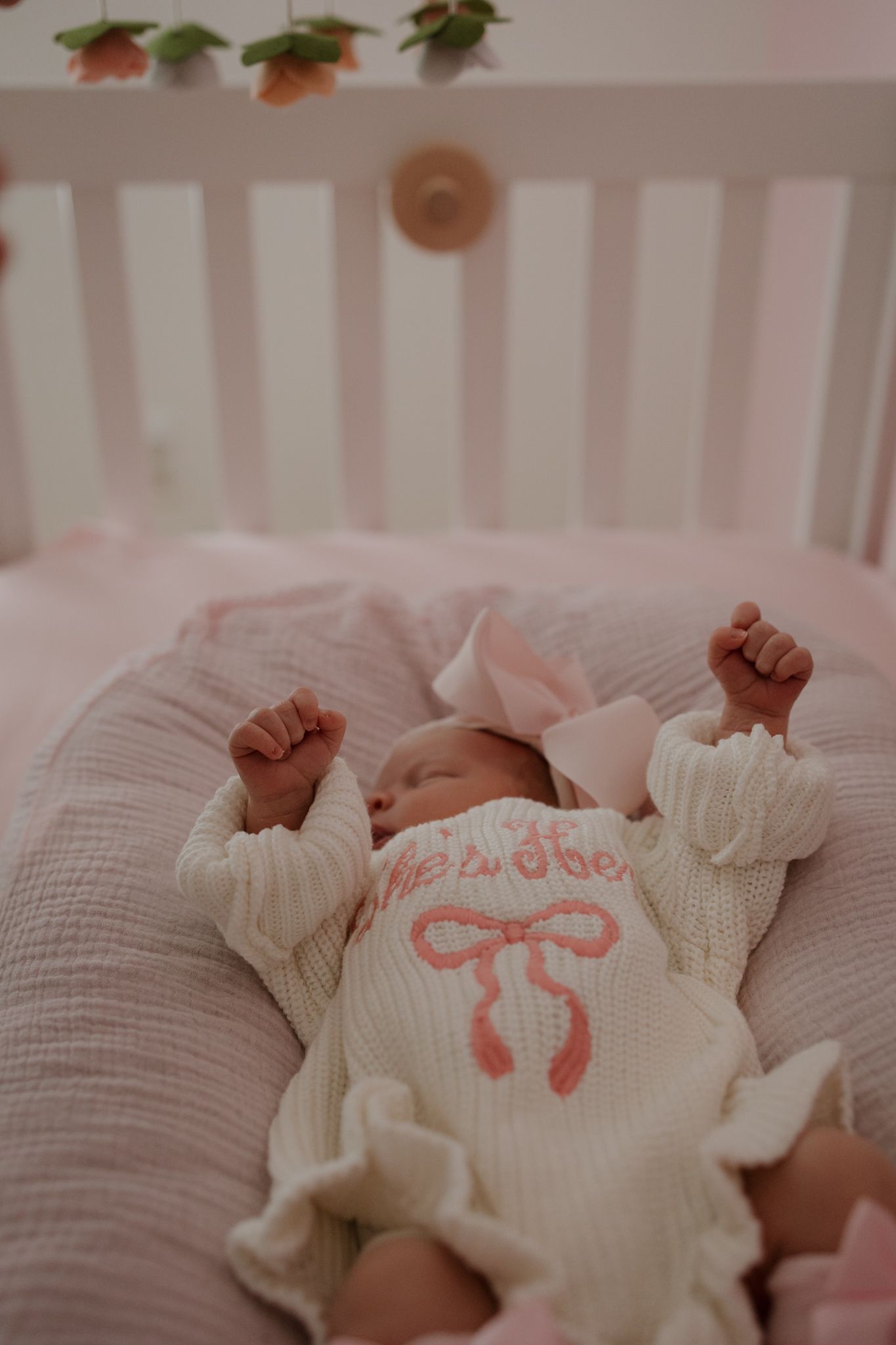 A sleeping baby in a white crib wearing a cream-colored outfit with a pink bow design stretches their arms upward.