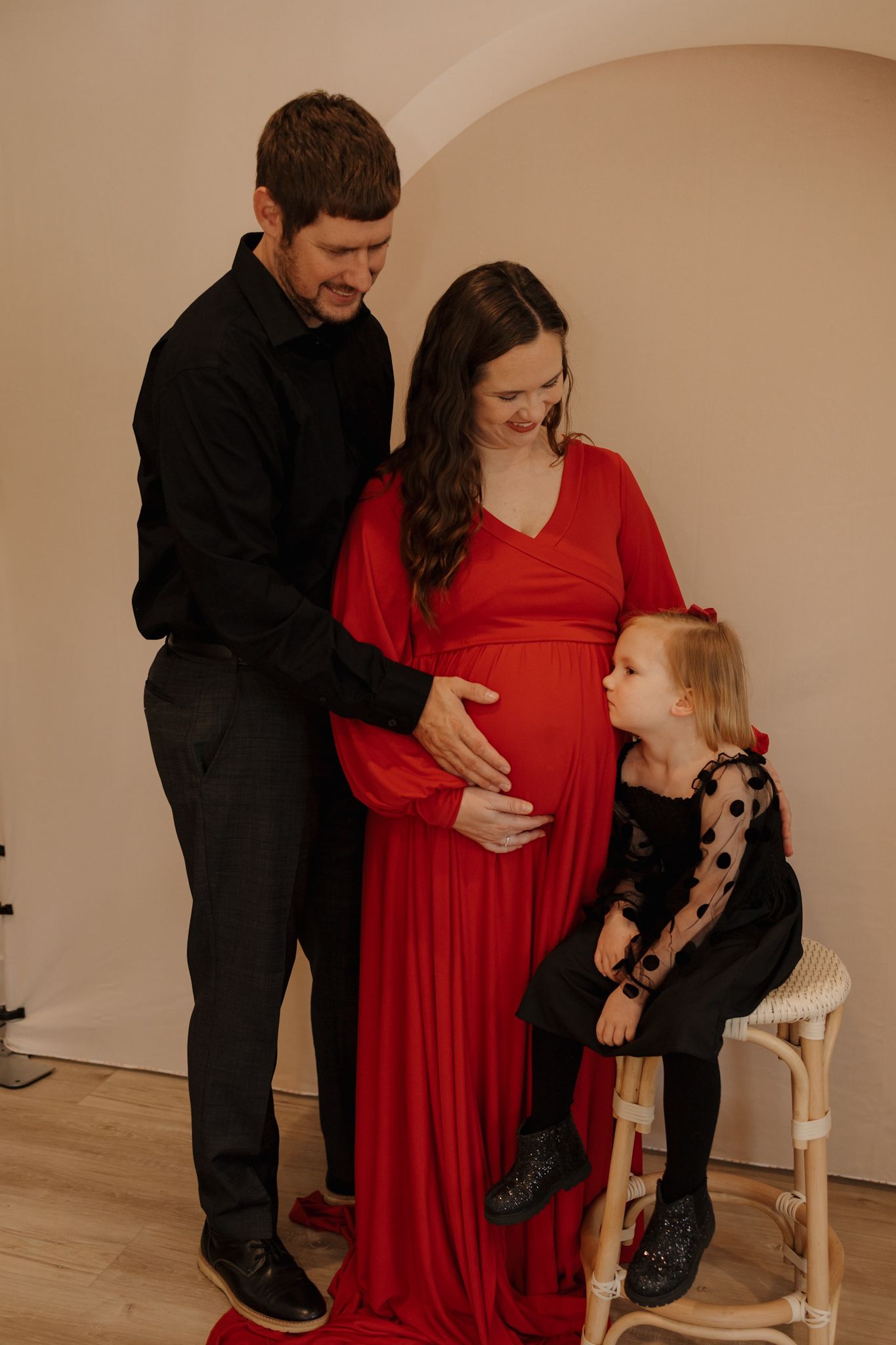 A couple in black and red attire poses with their small dog during a pregnancy photoshoot session.