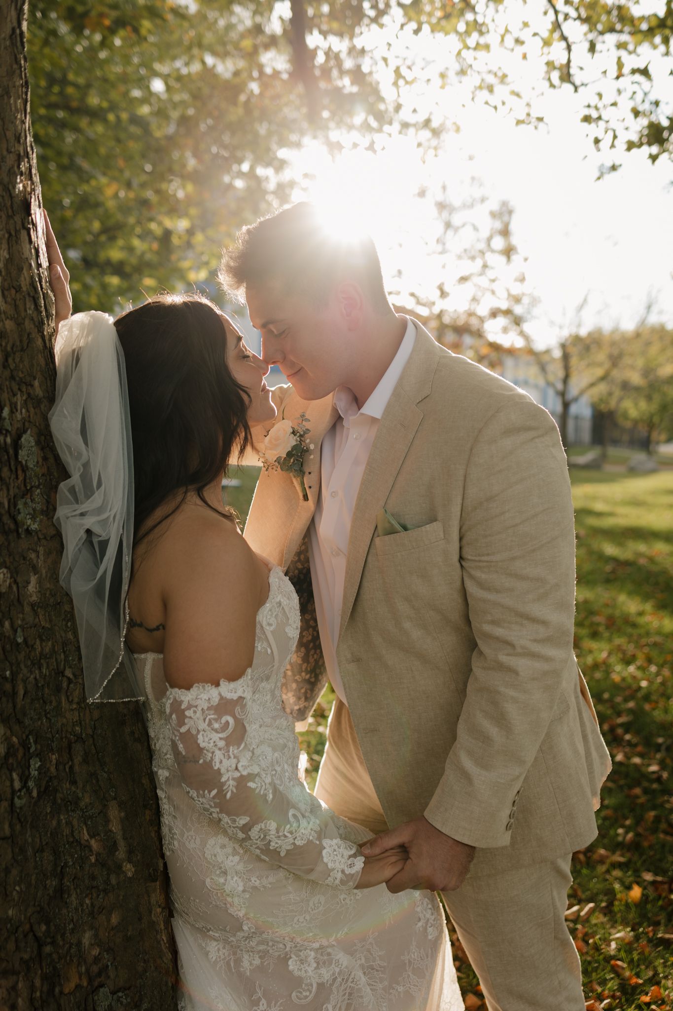 A romantic wedding couple shares intimate moments in a sunlit garden with autumn leaves on the ground.