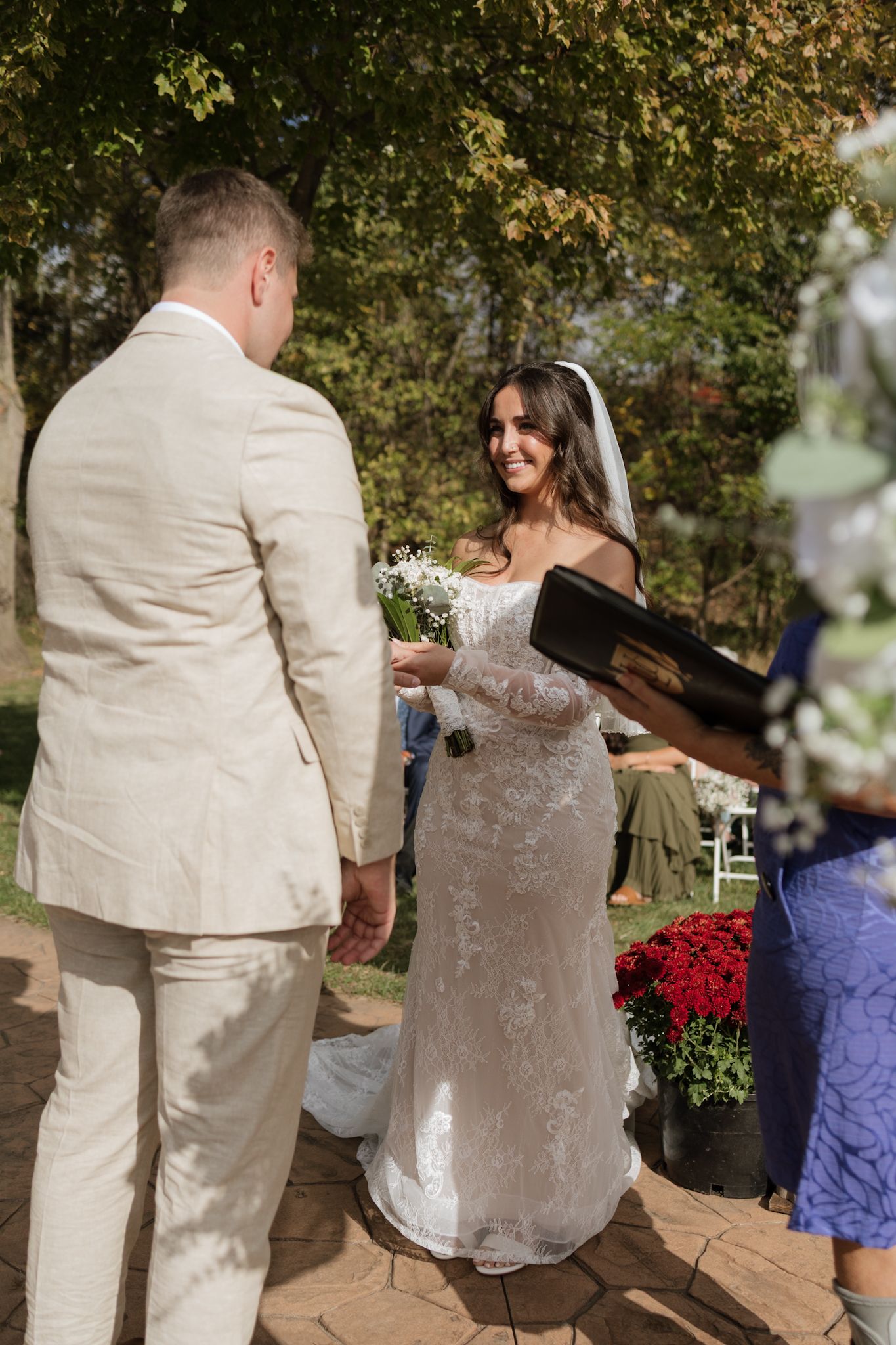 Wedding couple opens a champagne bottle in an outdoor garden setting with red flowers in the background.