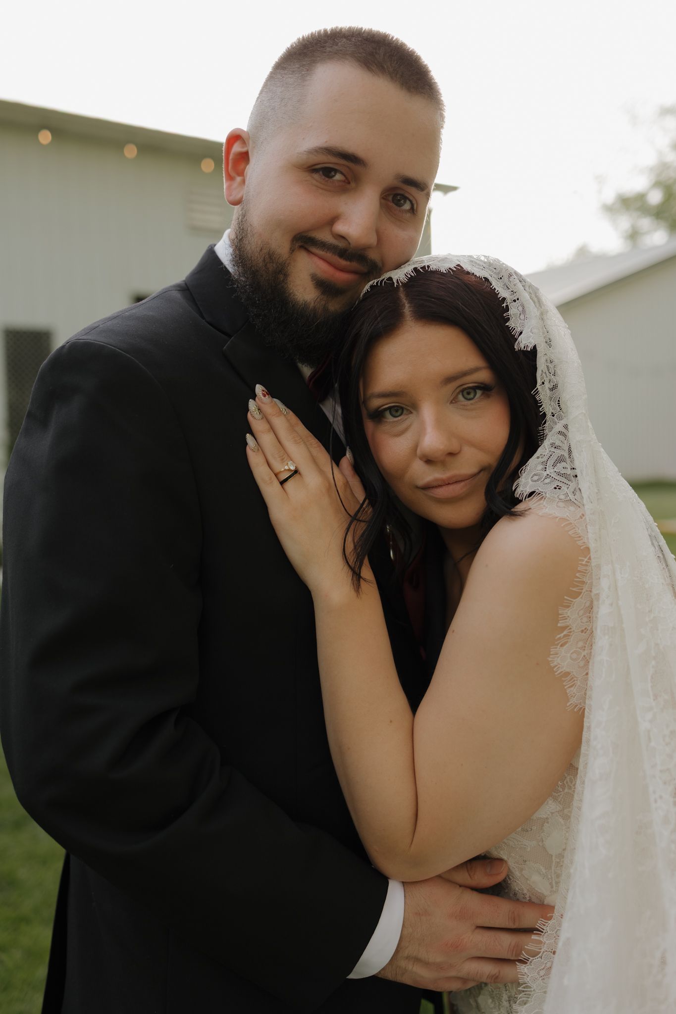 A bride and groom pose together outdoors, she wears a lace veil and he wears a black suit on their wedding day.